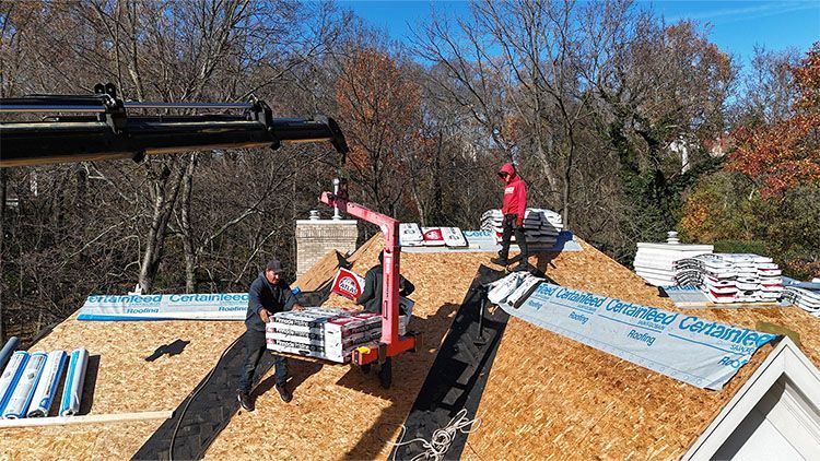 Roofing workers using a crane to lift shingles onto a house roof on a sunny day.
