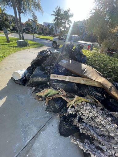 Piles of black garbage bags, cardboard, and yard waste stacked on a sidewalk near a driveway