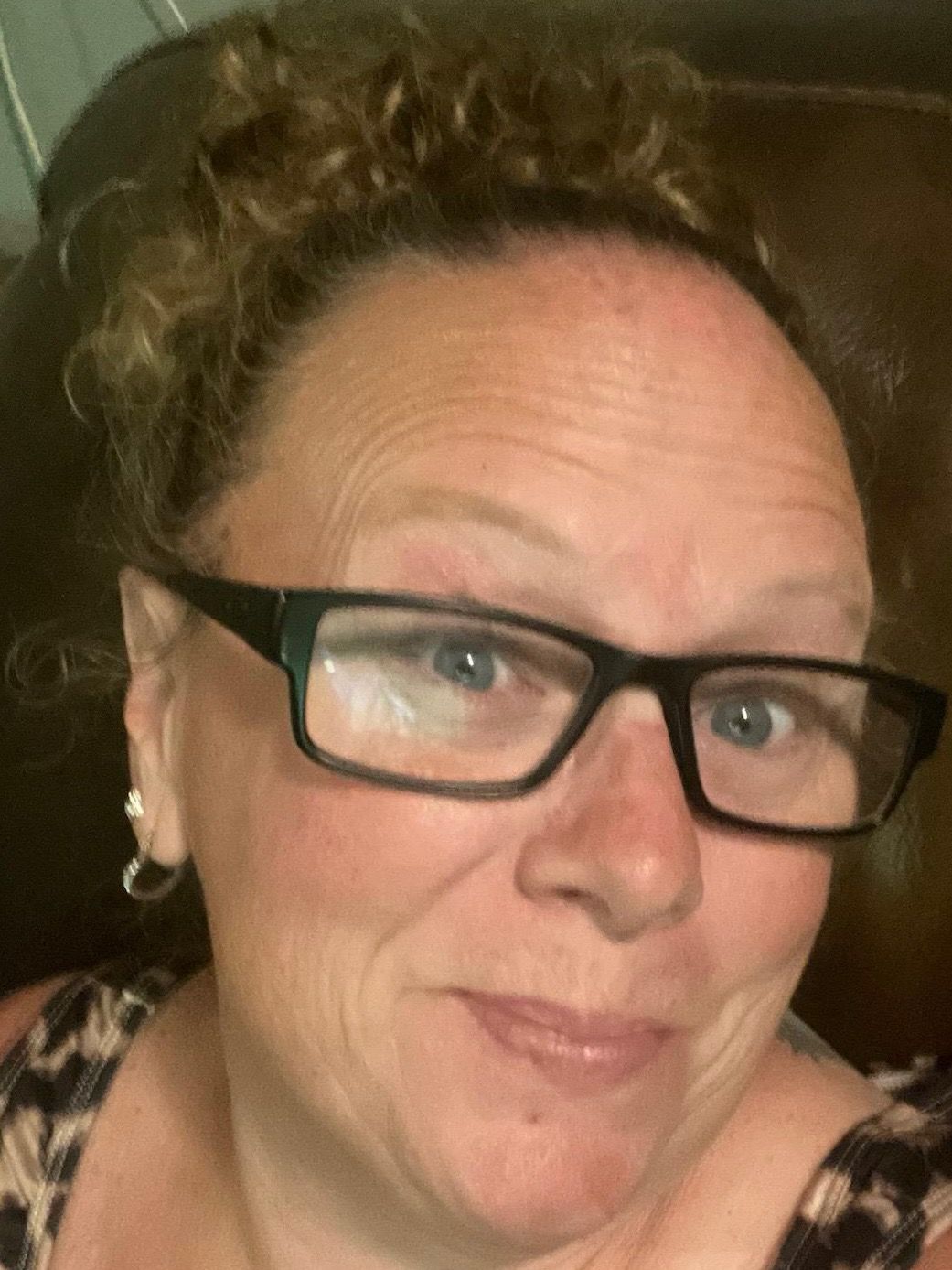 Woman with glasses, curly hair pulled back, smiling, wearing a patterned top.