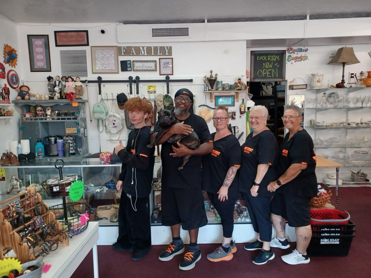 Five people, including one holding a dog, pose in a cluttered shop filled with merchandise. All wear matching shirts.
