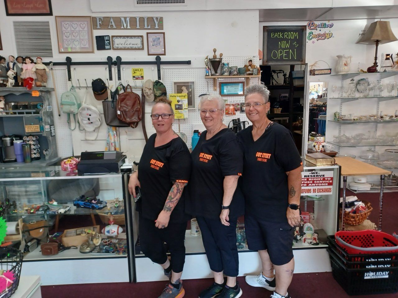 Three women in black shirts stand in a shop filled with merchandise.