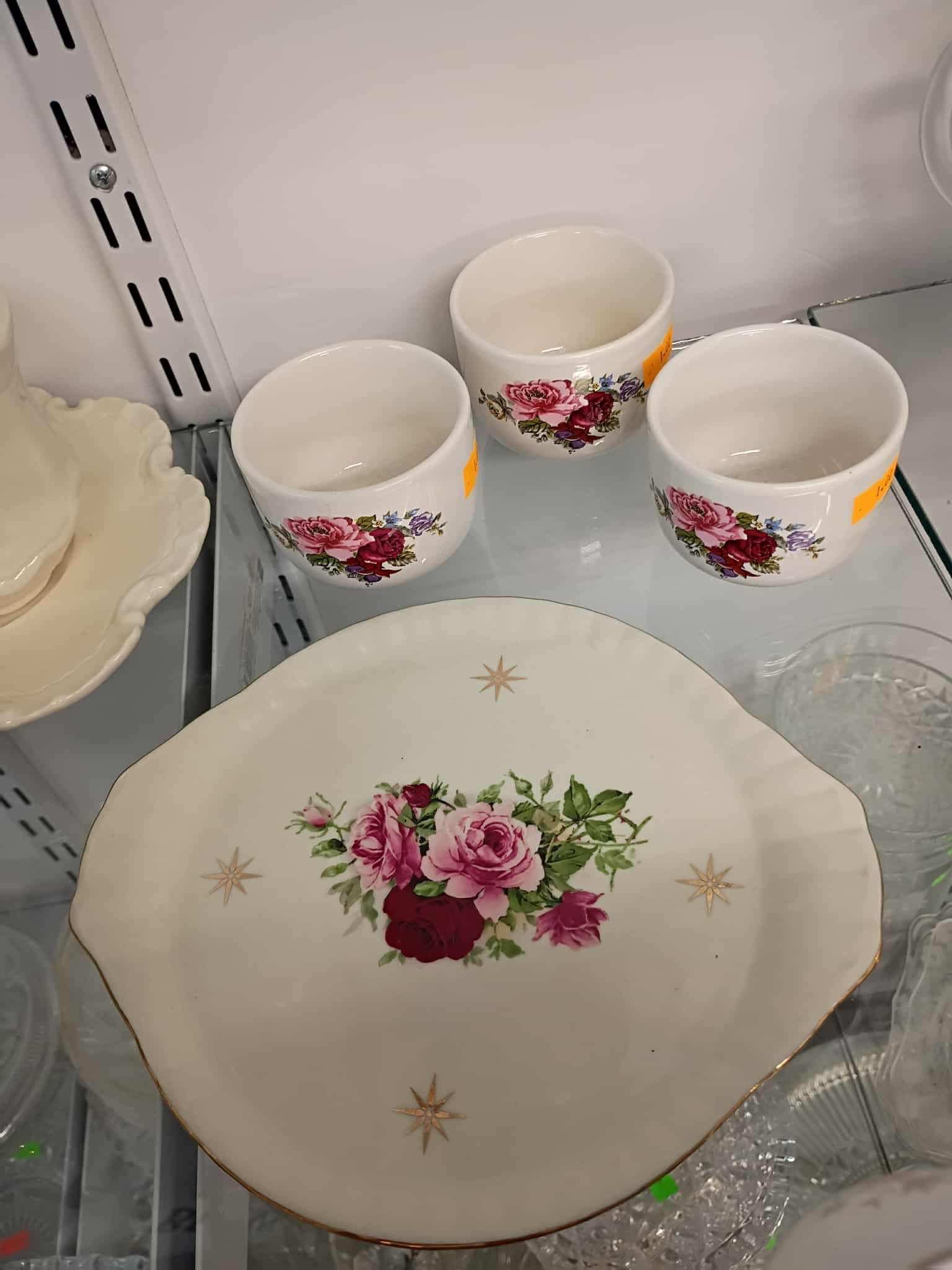 Three floral teacups and a matching platter with a rose design, on a glass shelf.