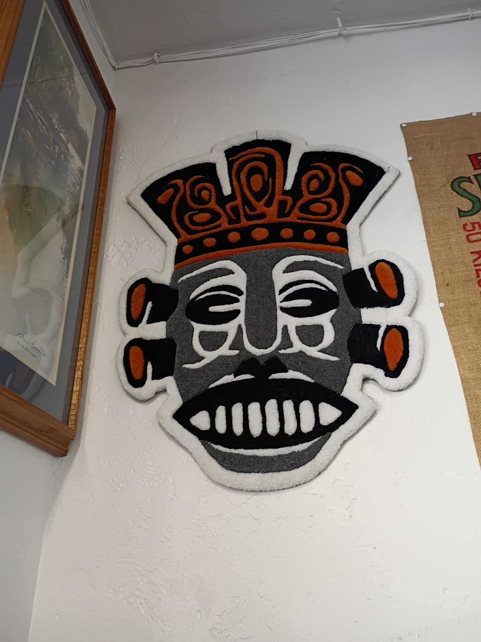 Mask-shaped rug with a smiling face, ornate crown, and side pieces. Mounted on a white wall.