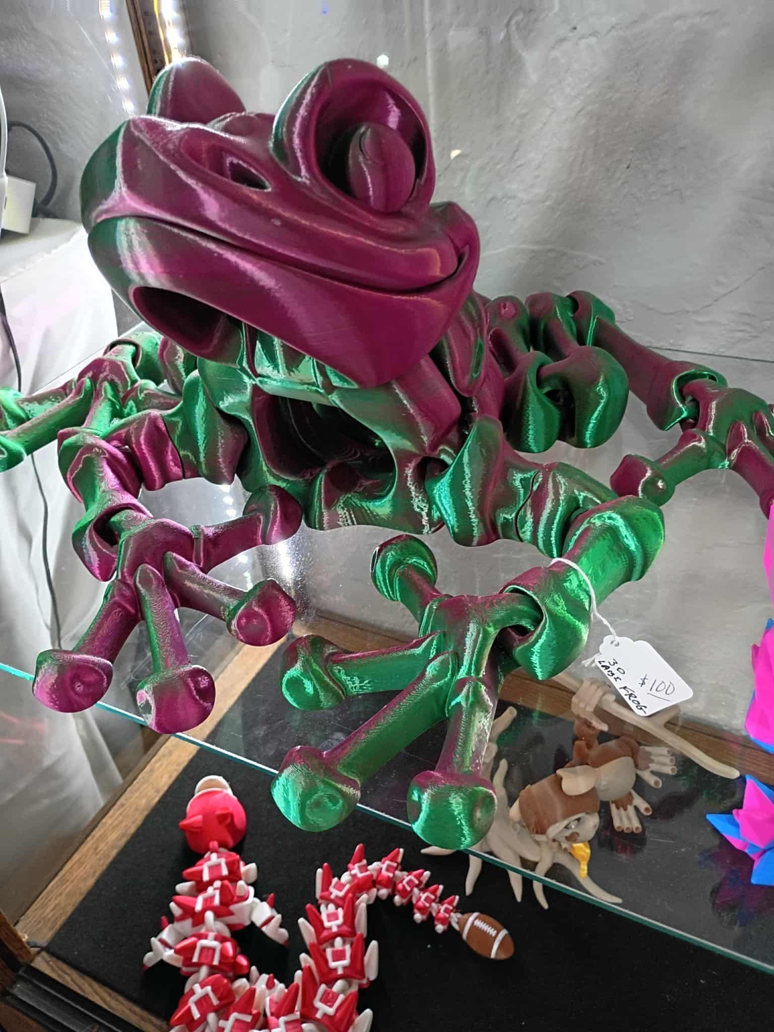 A 3D-printed articulating frog toy, green and purple, displayed in a glass case.