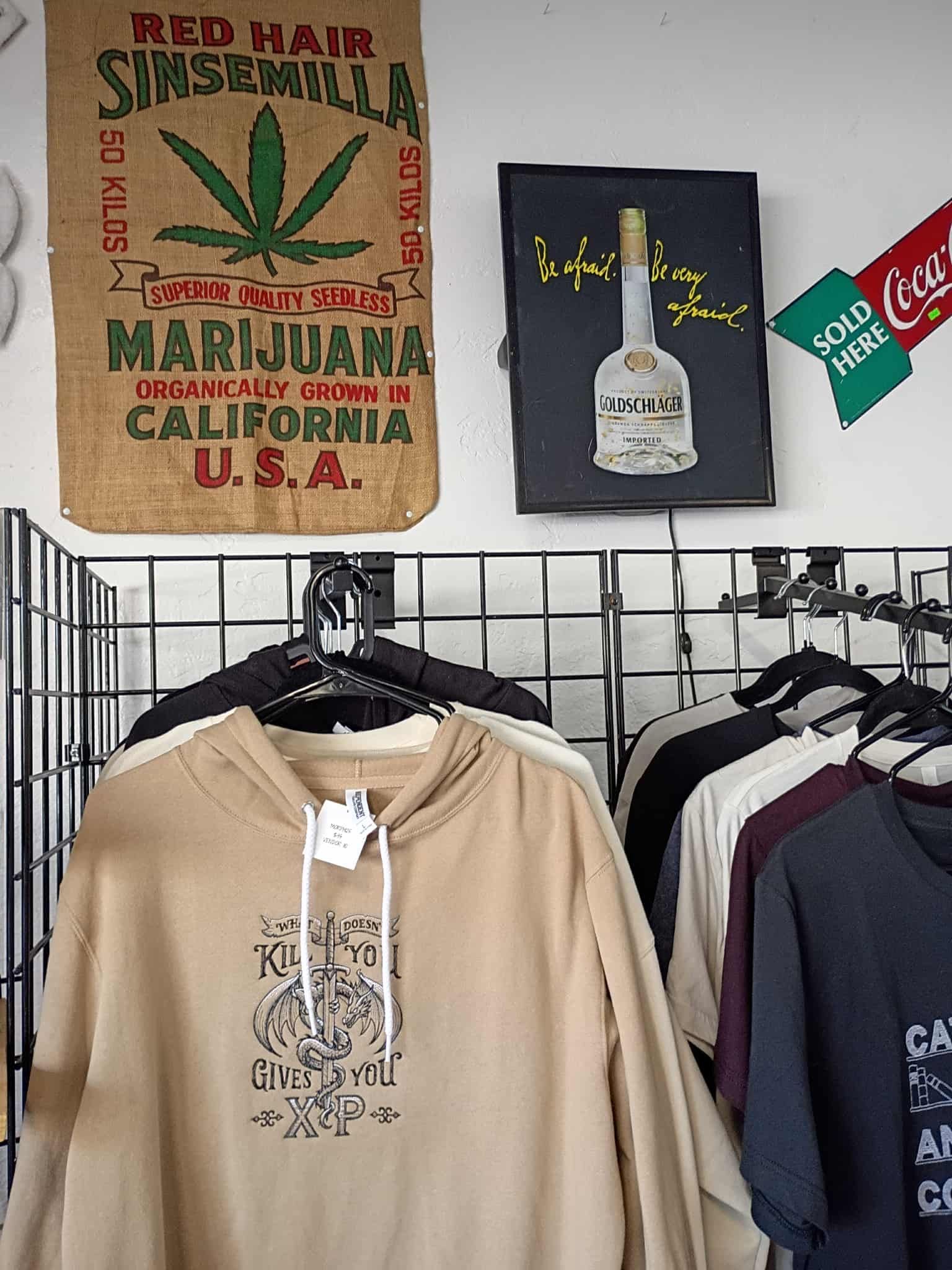 Clothing display with marijuana bag, artwork, and Coca-Cola sign. Hoodies and shirts hang below.