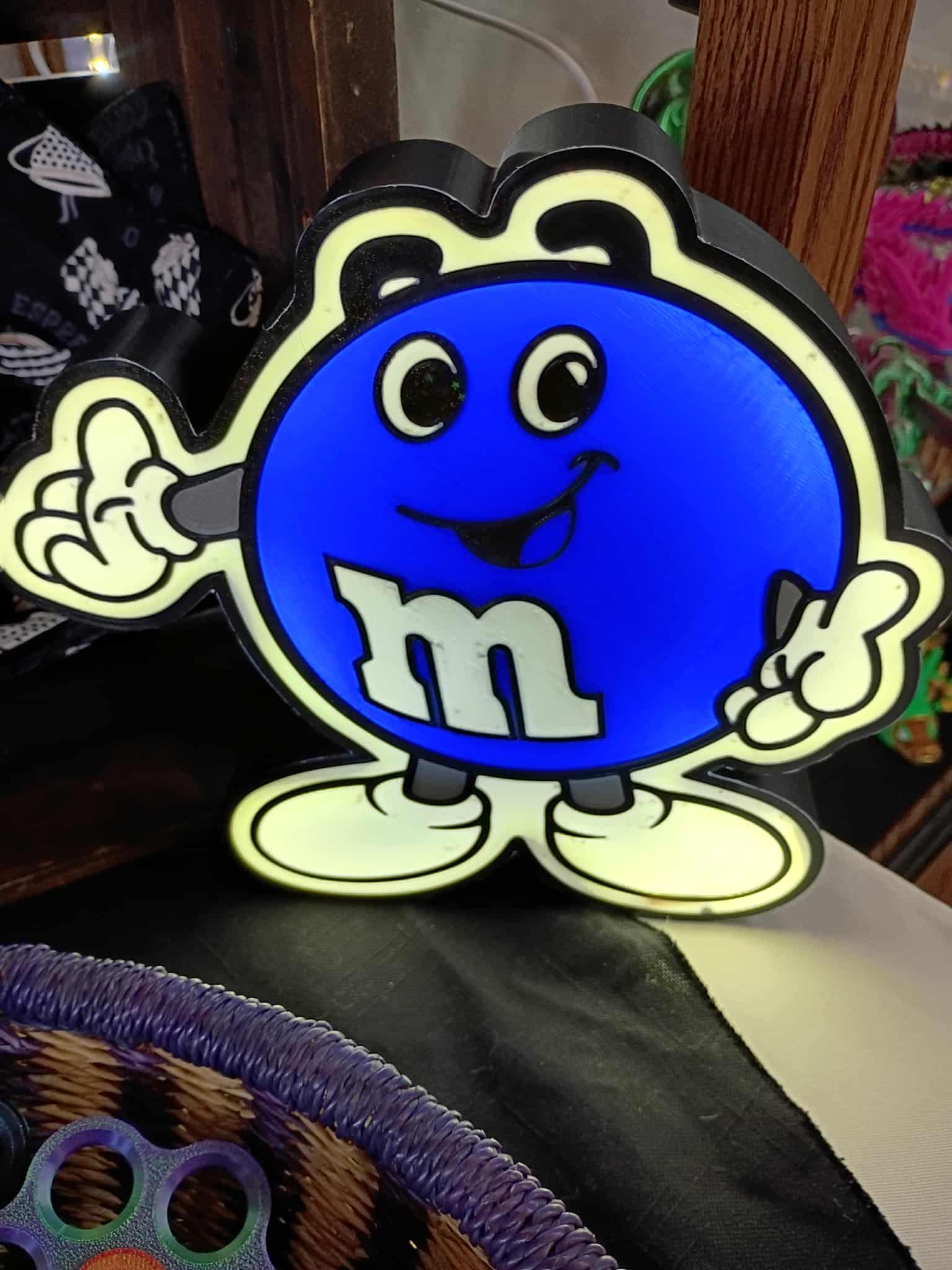 Blue M&M character with white outlines, smiling and waving.