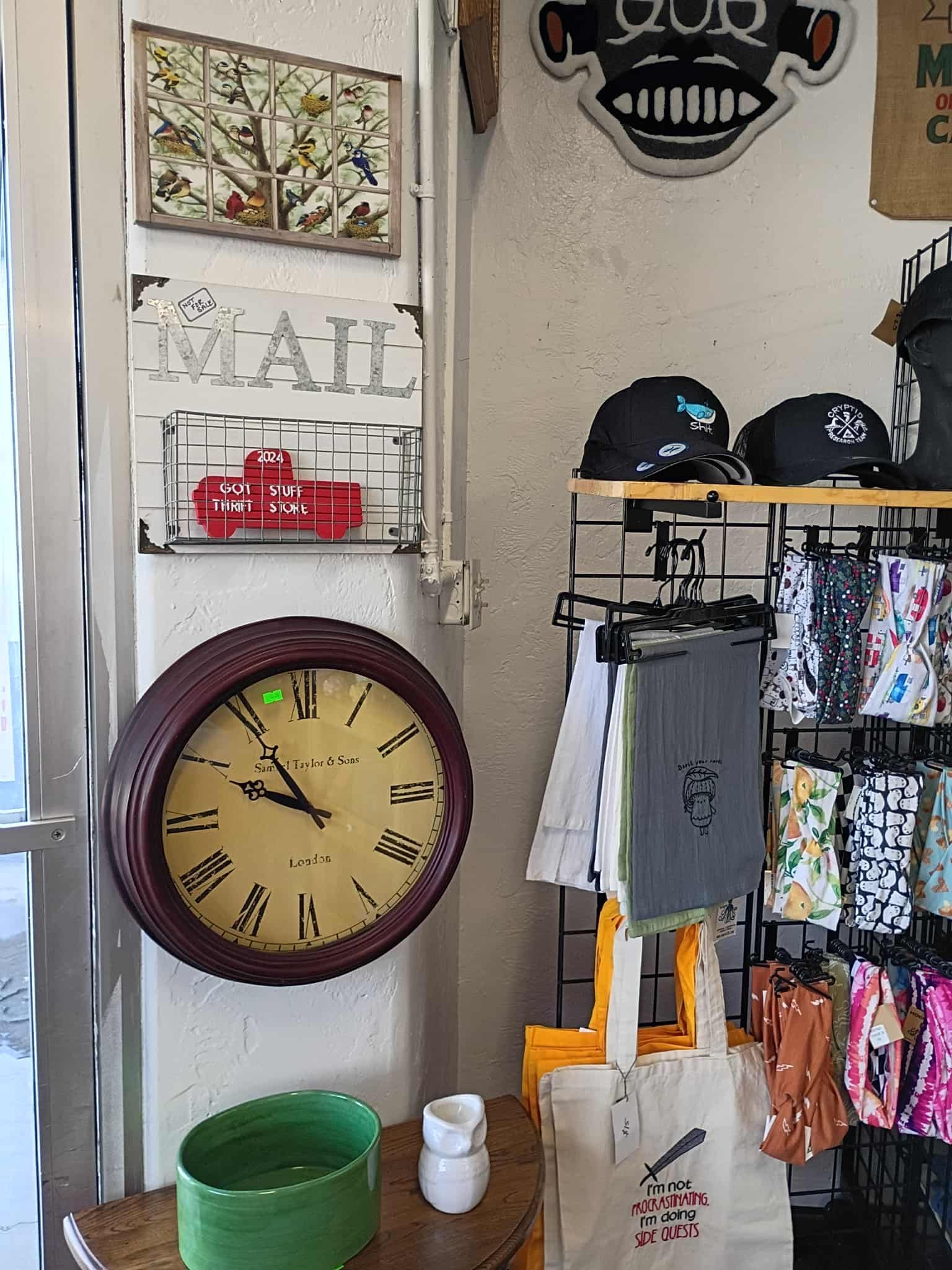 Shop interior with clock, mail rack, and merchandise, including hats and tote bags.