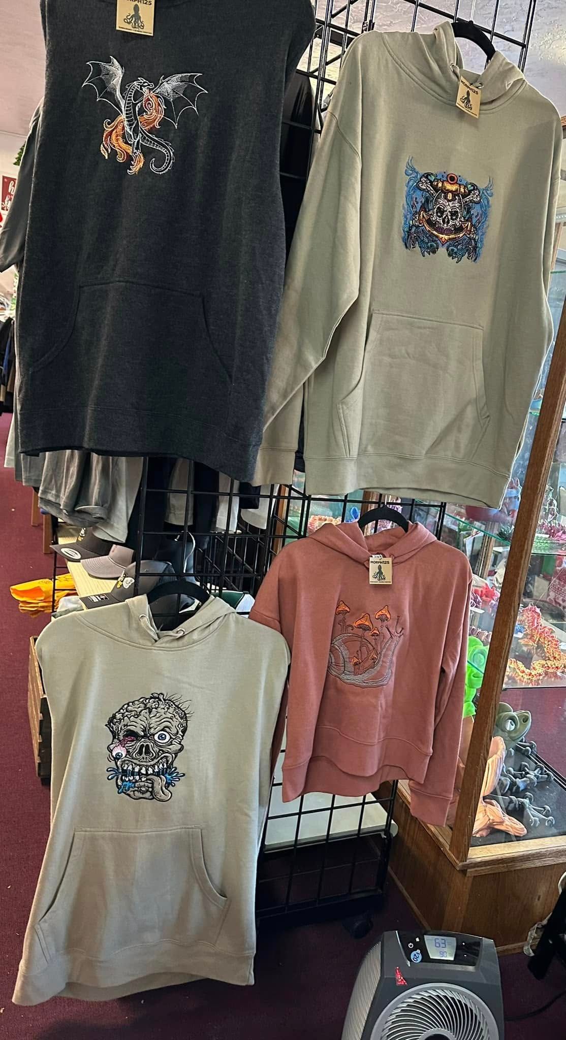 Apparel display: four hoodies with floral designs, in gray, green, pink, and cream, hanging on a rack.