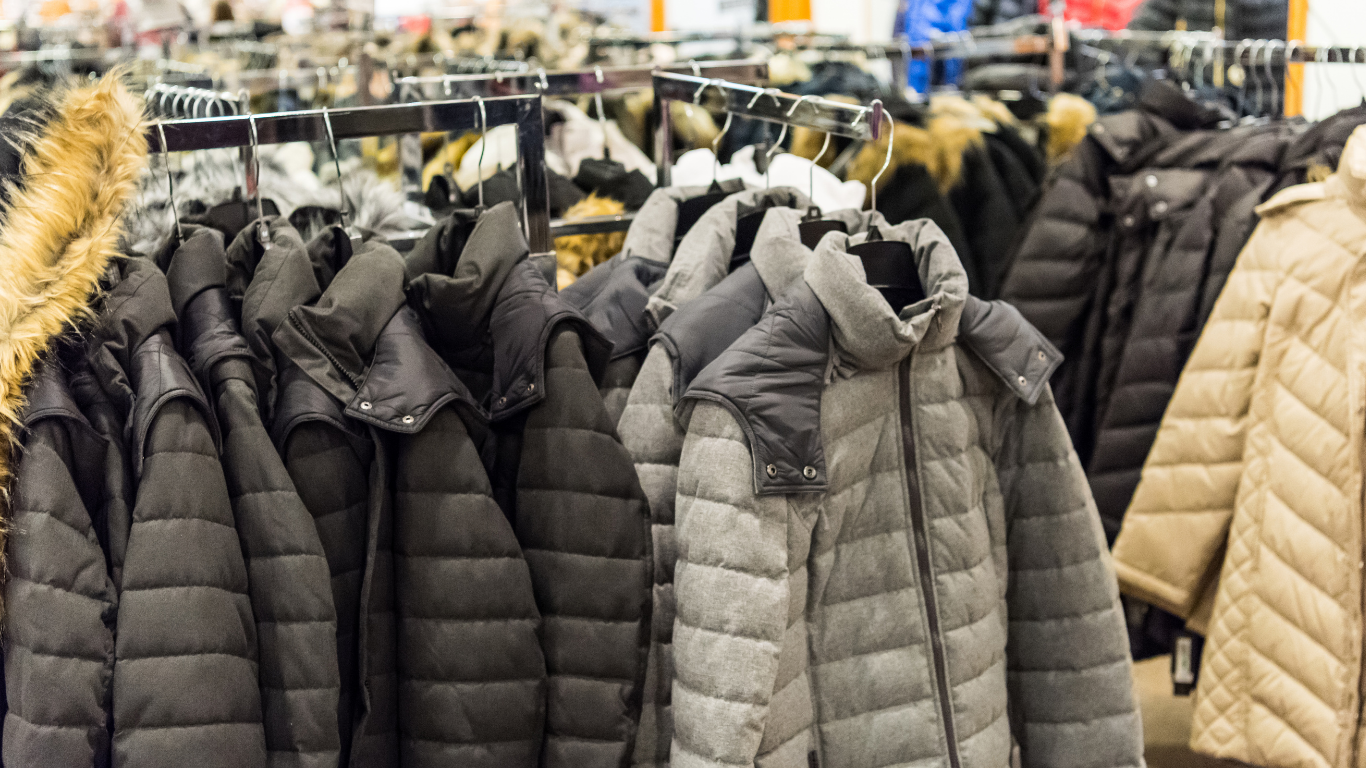 Rows of winter jackets hanging on a clothing rack in a store, mostly black and gray, with hoods.