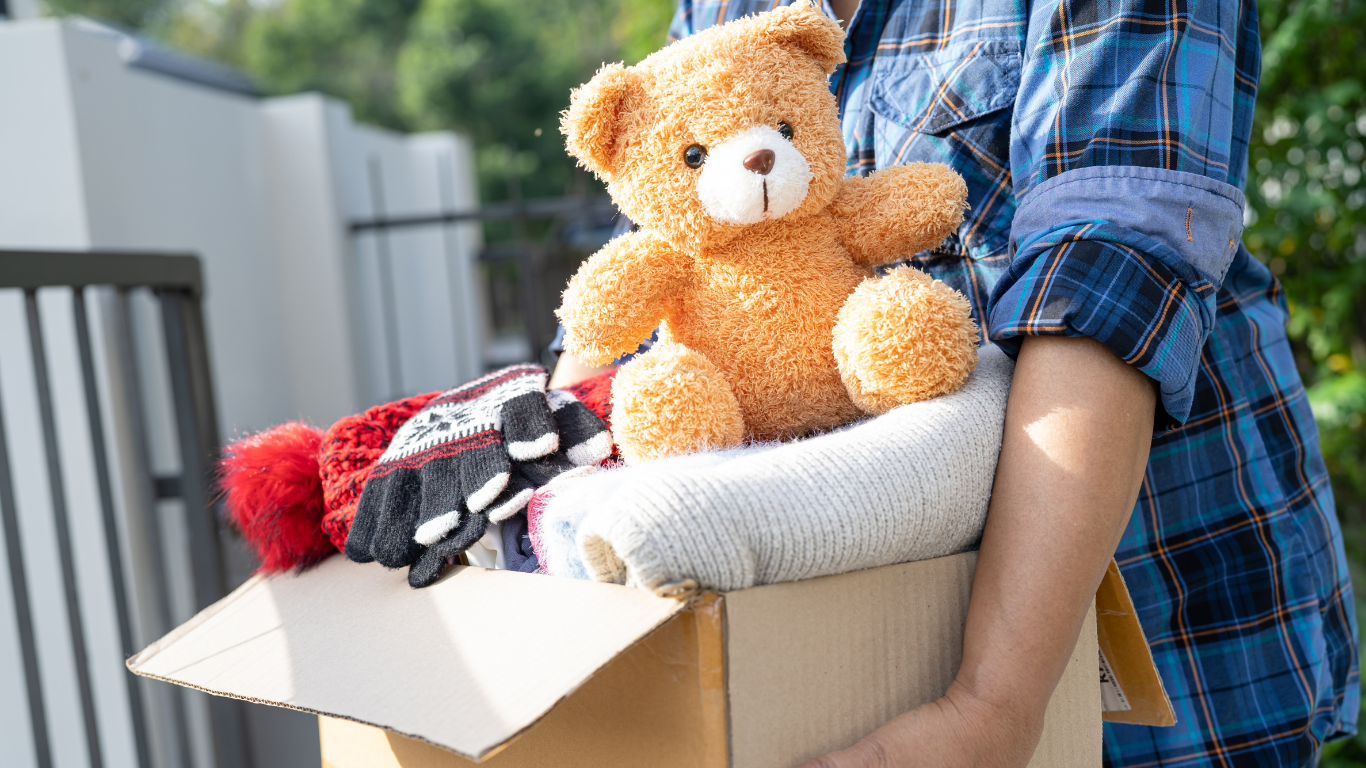 Person holding a cardboard box filled with a teddy bear, gloves, and clothing; outdoors.