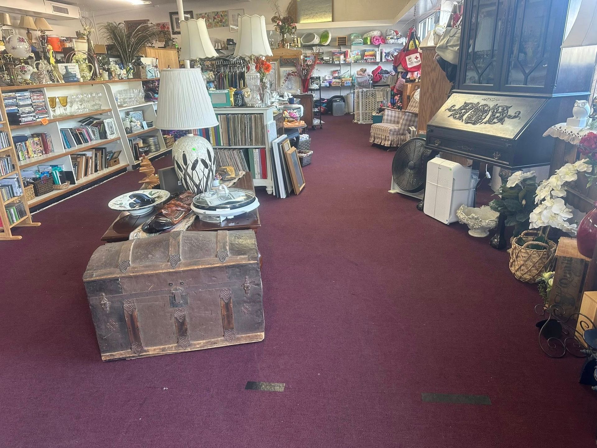 Antique shop interior, featuring shelves of items, furniture, and an old wooden chest on a burgundy carpet.