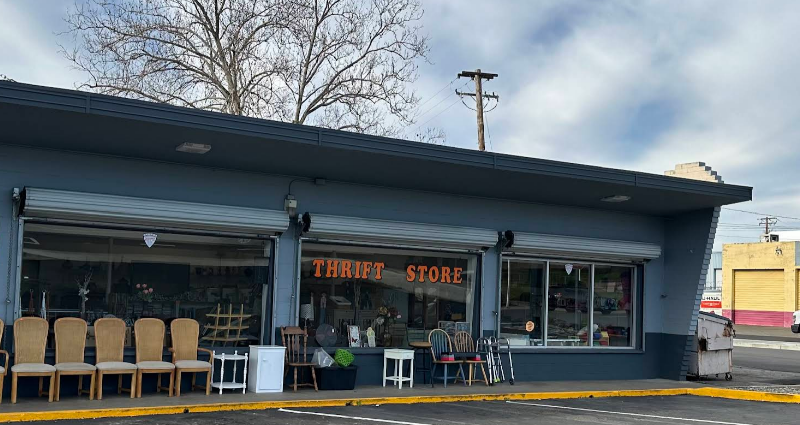 Thrift store exterior with chairs, a sign, and merchandise visible through windows. Gray building under cloudy sky.