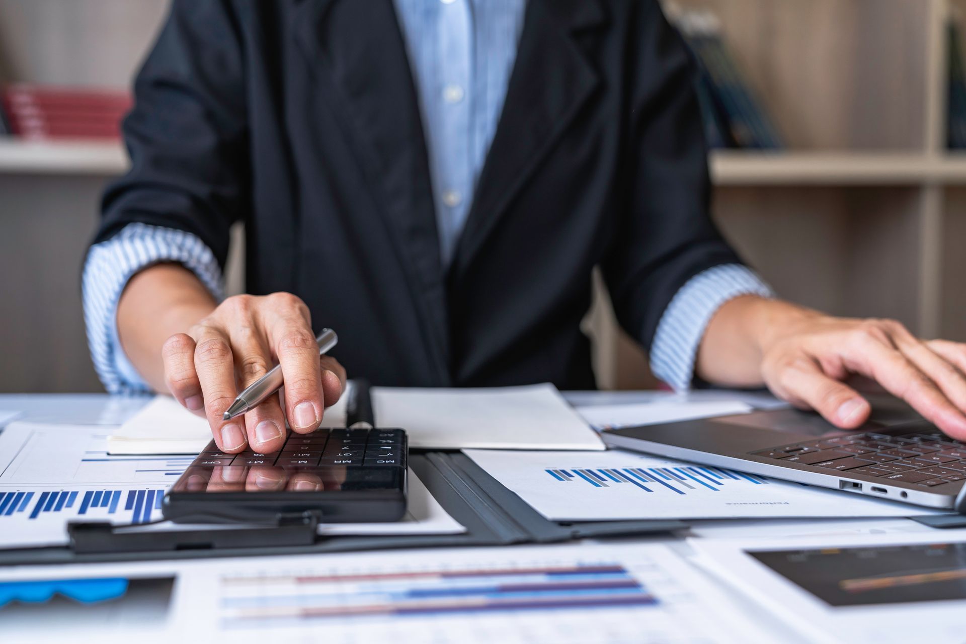 Close-up of hands using a calculator and reviewing documents next to a laptop.