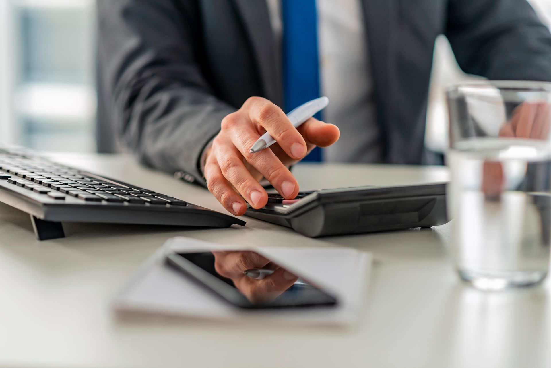 Close-up of an accountant as they use a calculator, a keyboard, and a phone, on a white table.