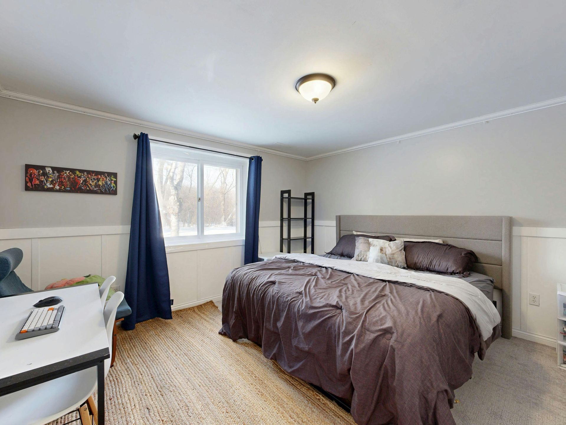 Bedroom with bed, desk, window, and rug. Gray walls, dark blue curtains, and light-colored bedspread.