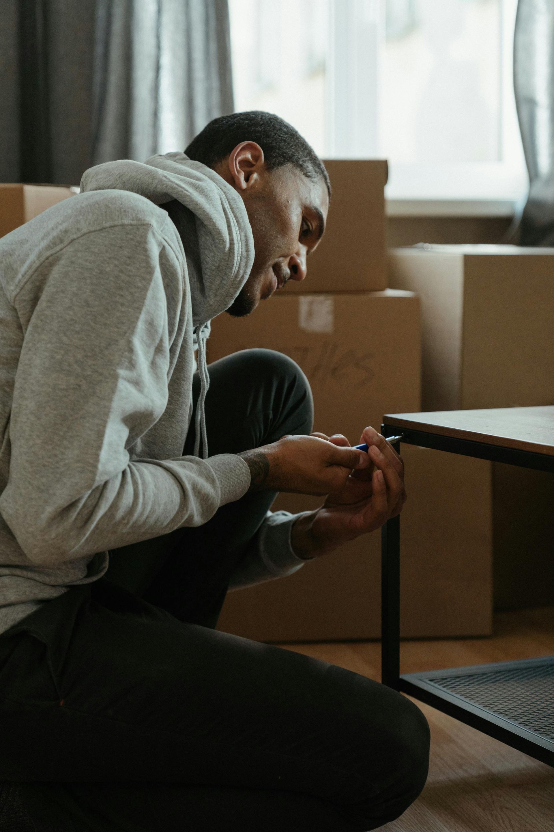 Person in gray hoodie assembling furniture, kneeling, with moving boxes in background.