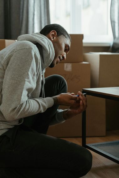 Person in gray hoodie assembling furniture, kneeling, with moving boxes in background.