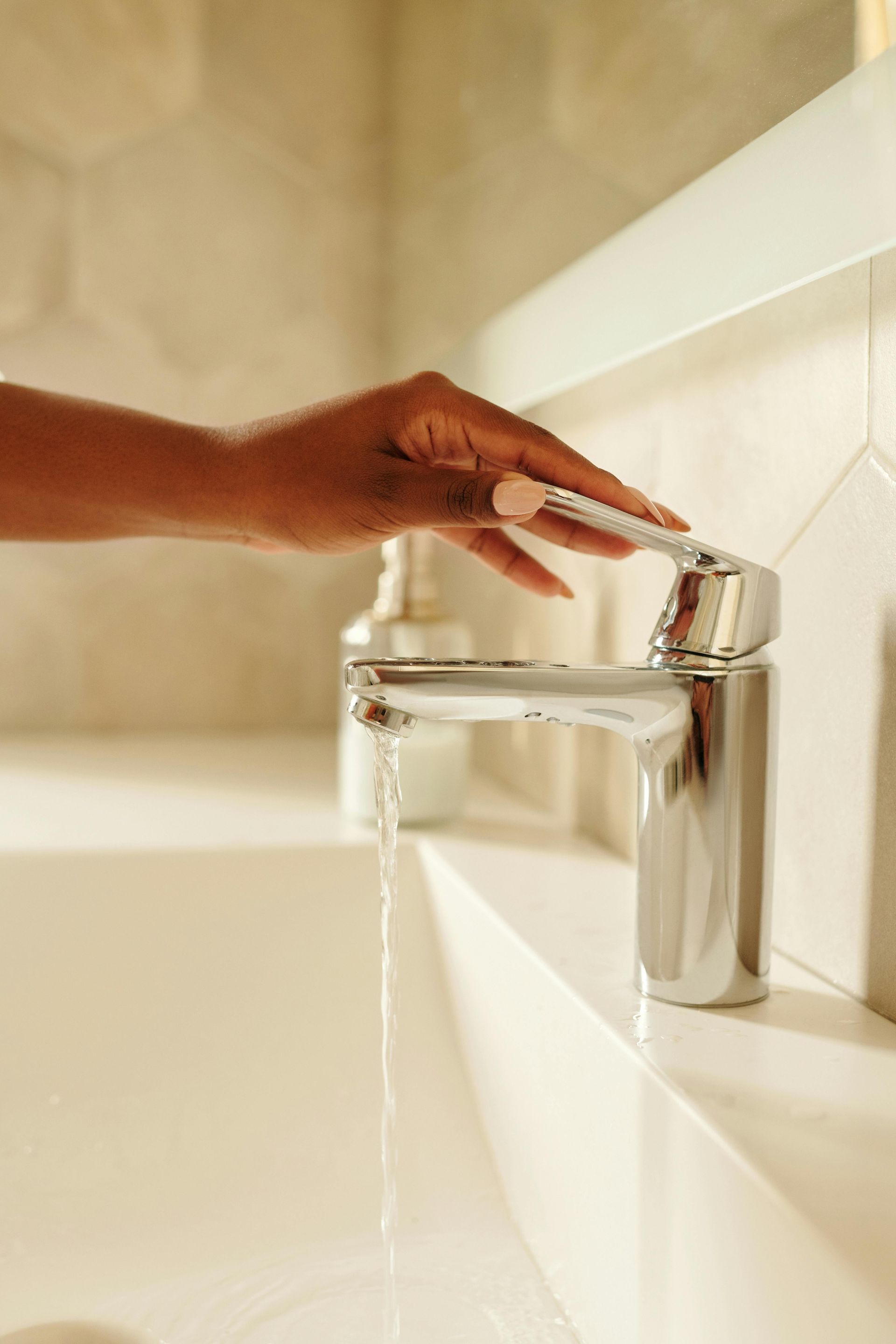 Hand turning on a chrome faucet in a bathroom, water flowing into the white sink.