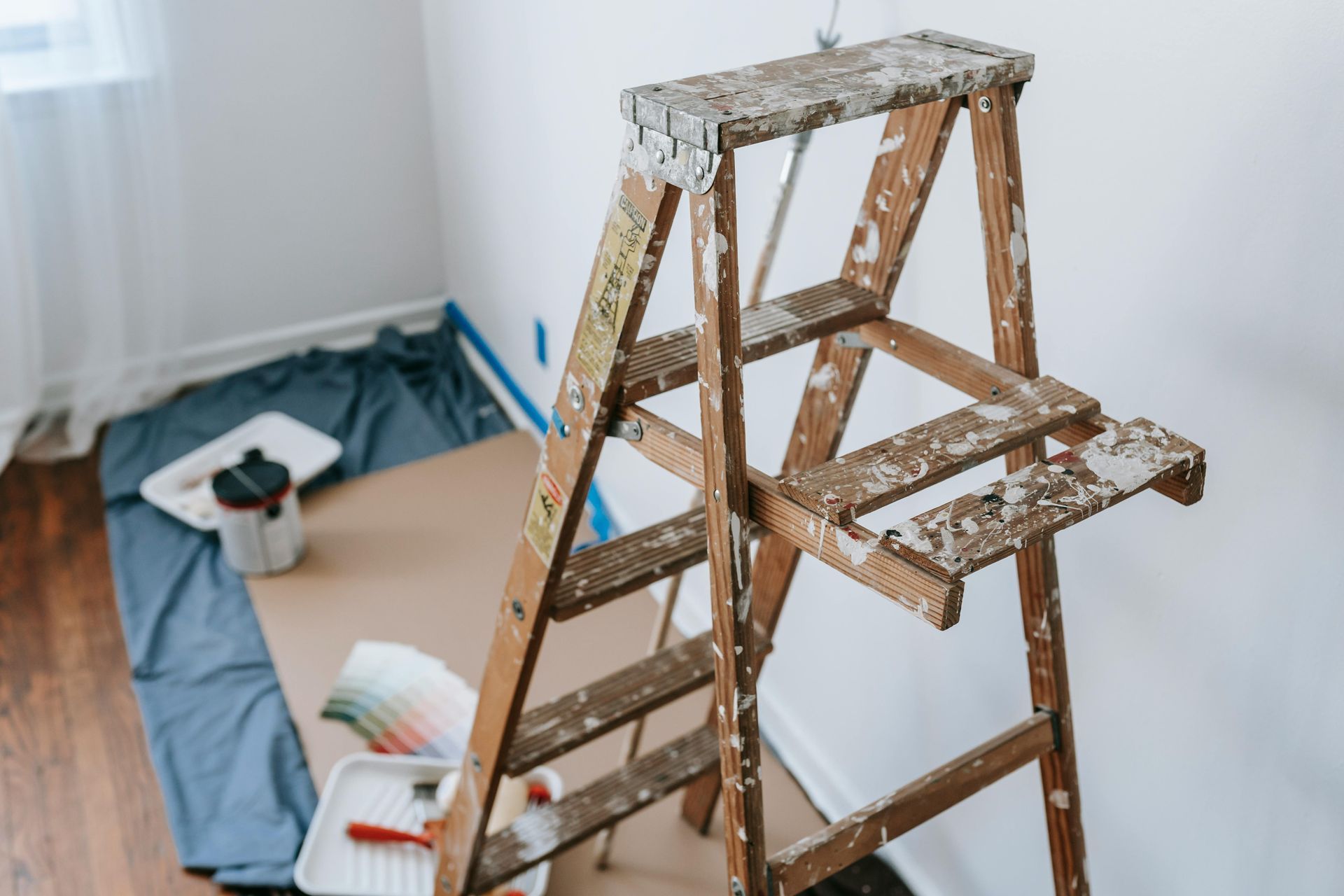 Wooden step ladder with paint splatters, near paint supplies and a tarp in a room being painted.