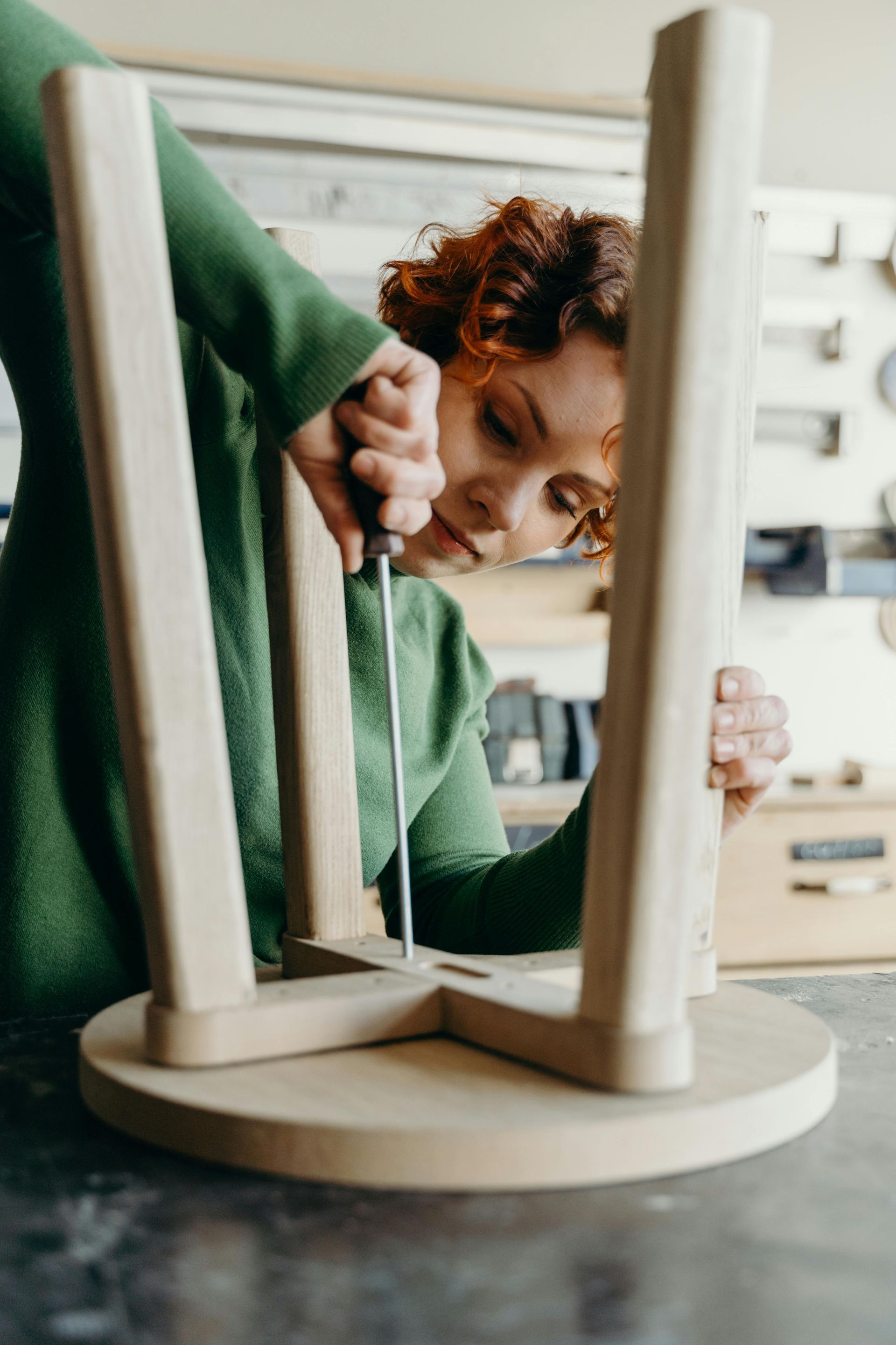 Person in green sweater assembles wooden stool with a screwdriver in a workshop.