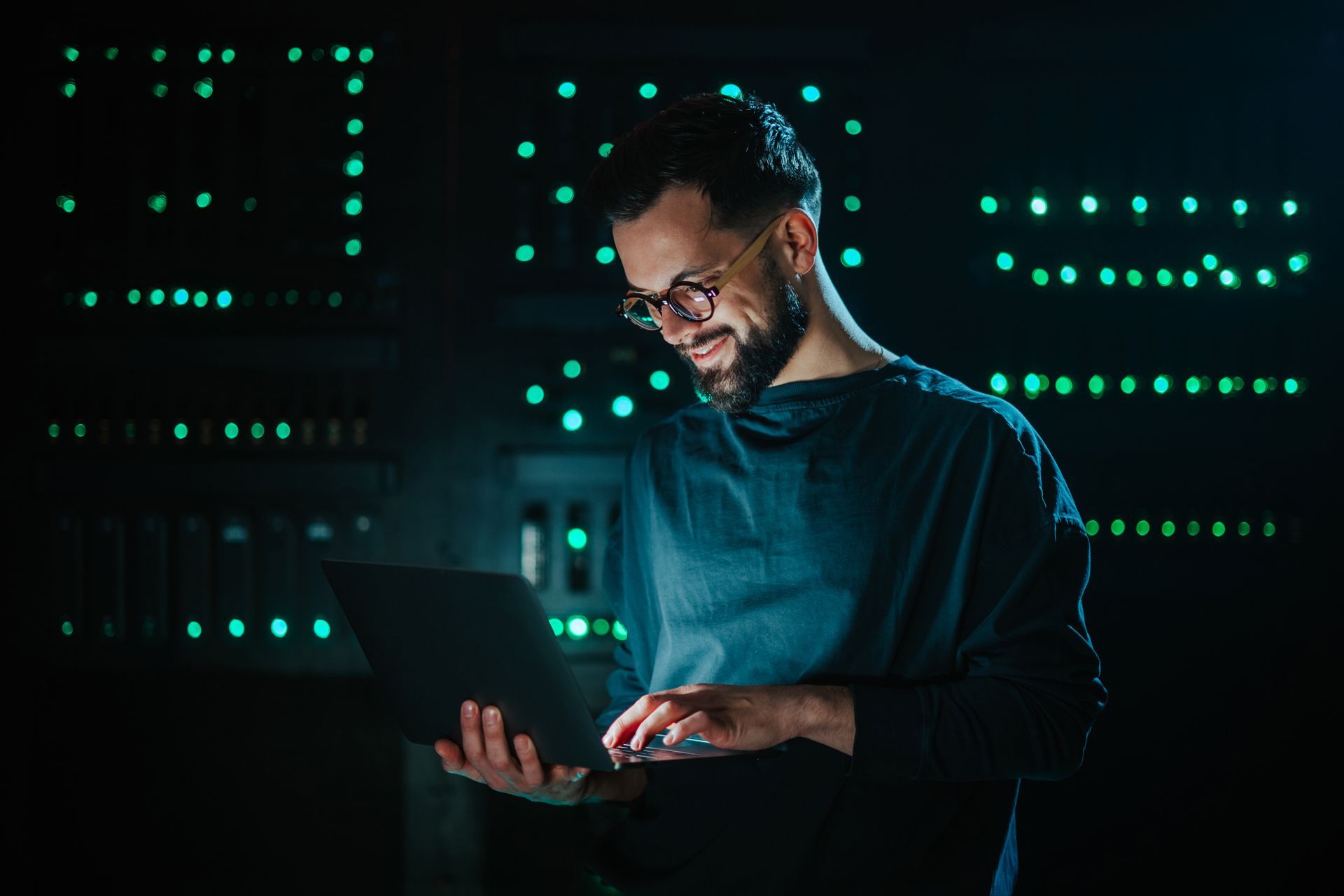 An engineer holding a laptop working on a server in a dark, green-lighted data centre.