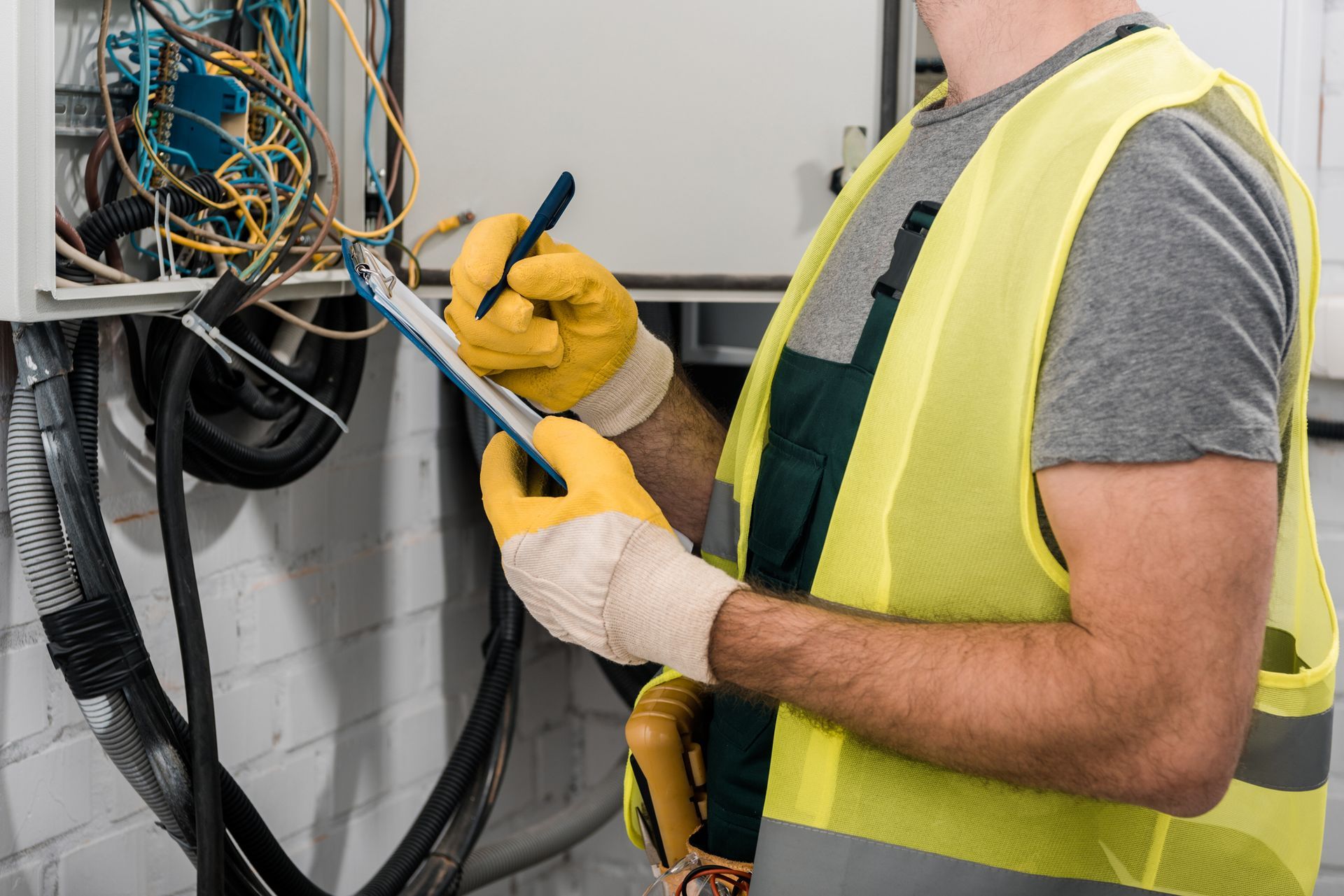 A male electrician holds a clipboard and pen near an electrical box with cables, inside a corridor.