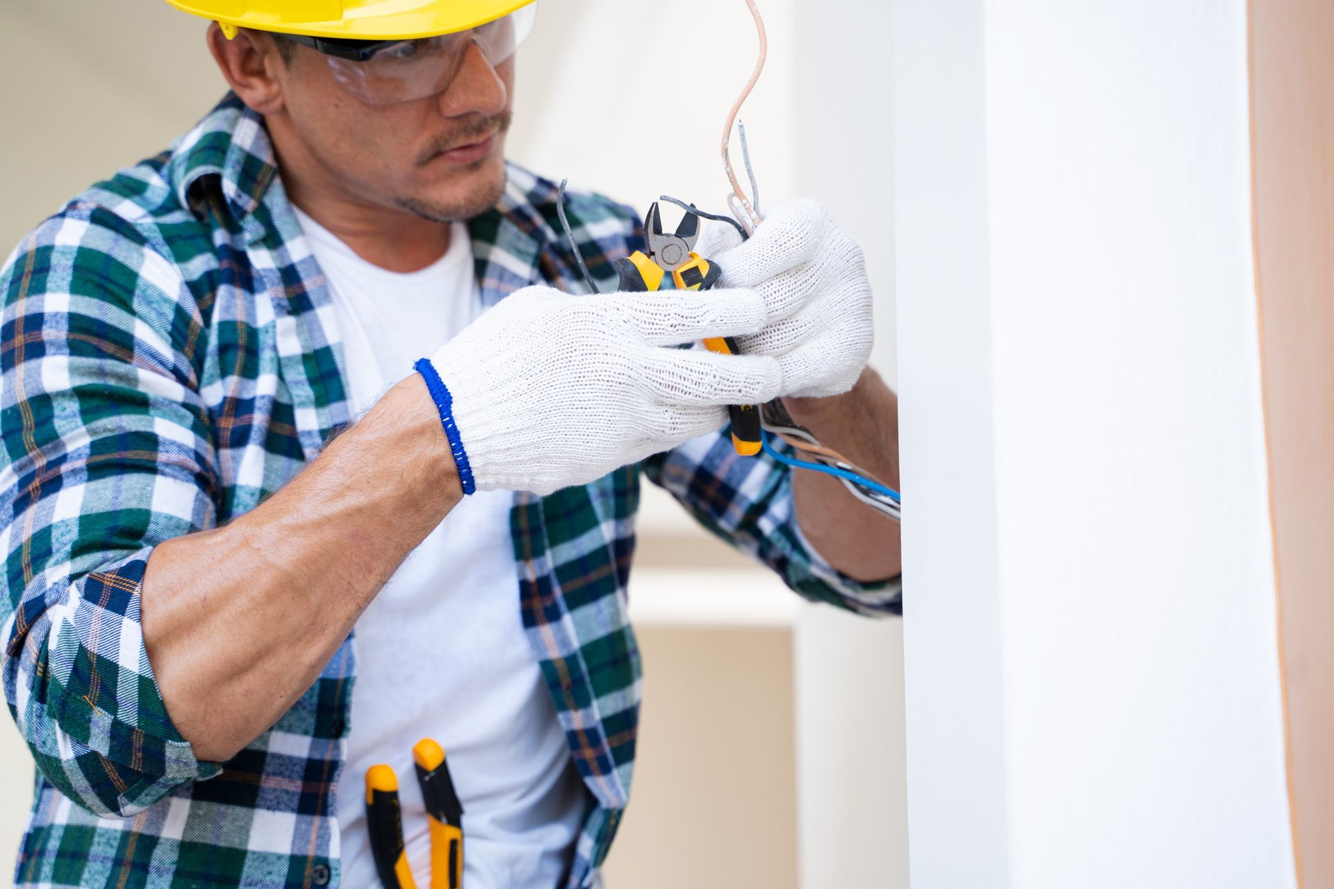 Electrician in a plaid shirt and hardhat, using wire cutters on wires.