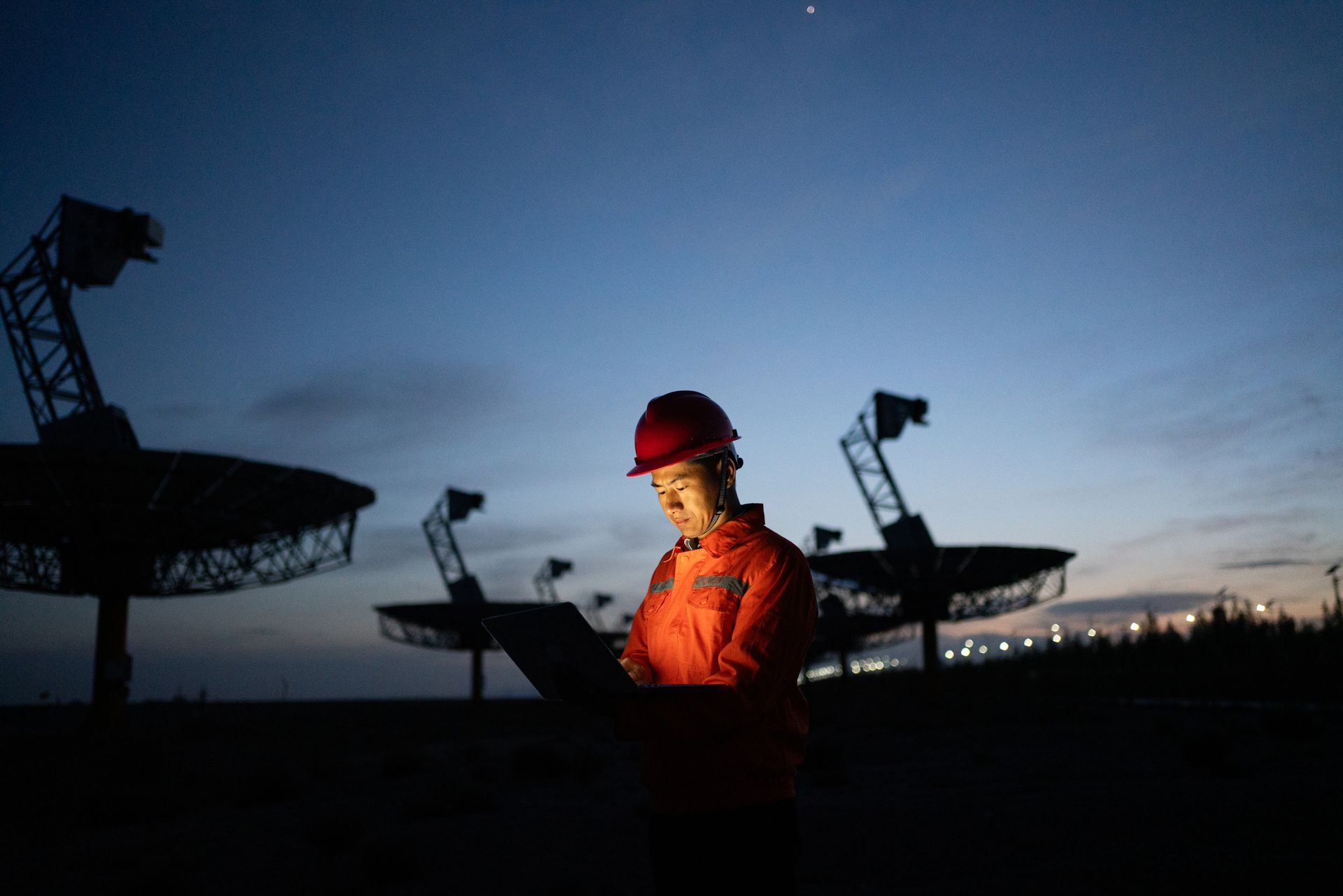 A person in an orange jumpsuit and hard hat works on a laptop at night, in front of several radio telescopes under a dark blue sky.