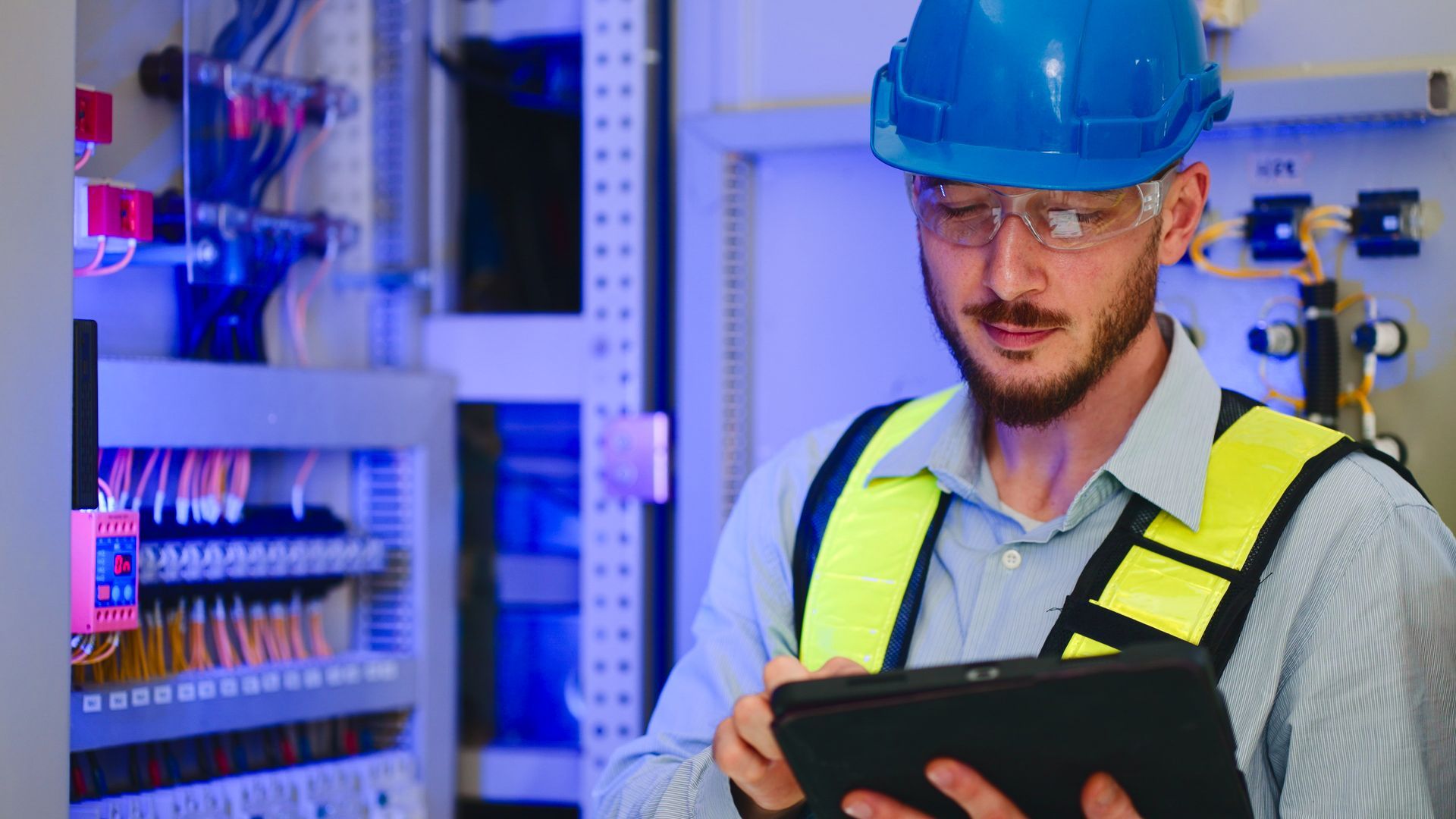 Electrician in safety gear using tablet near electrical panel.