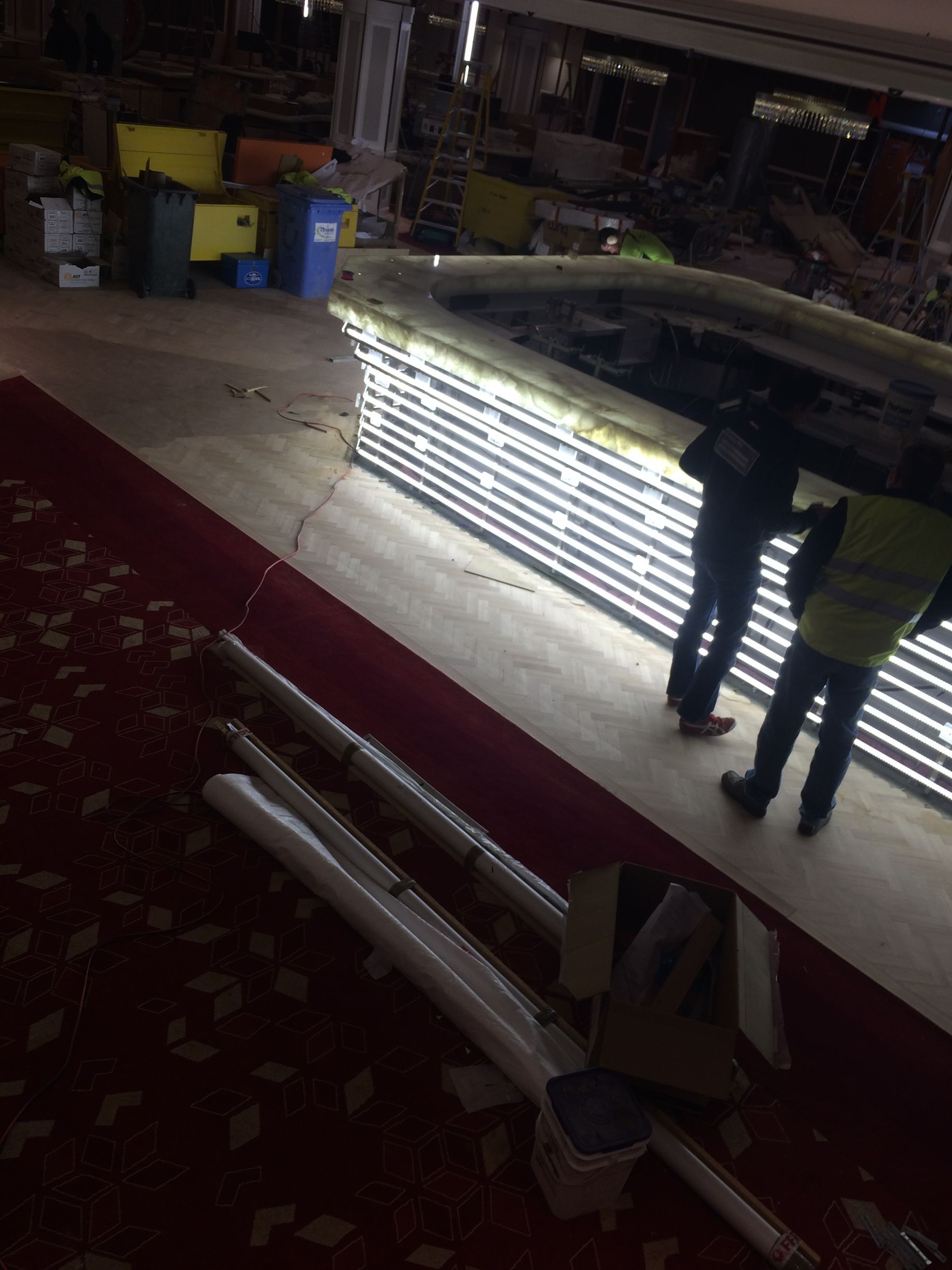 Construction site with workers near a long, illuminated bar. Red carpet and construction materials are on the floor.