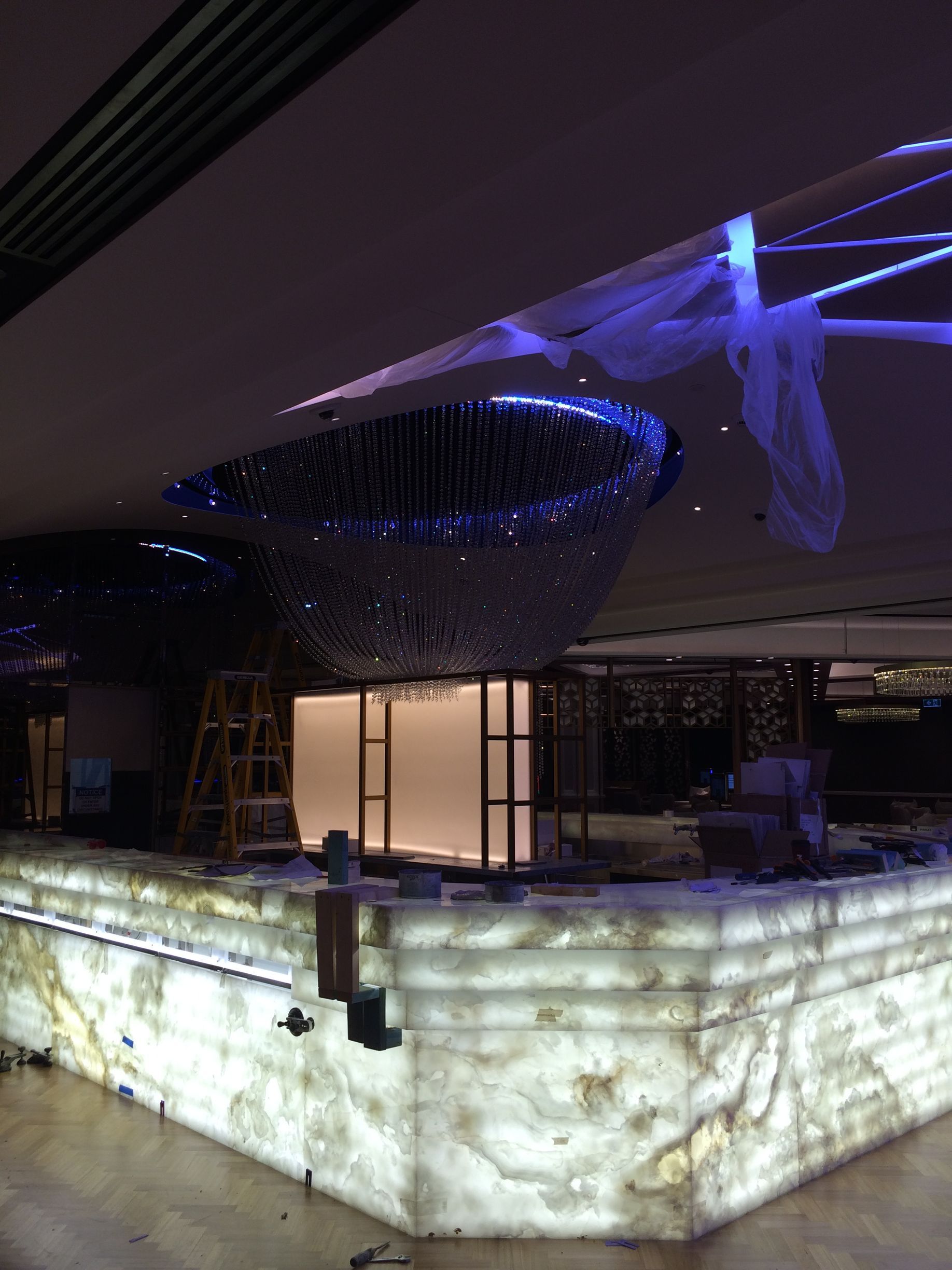 A bar under construction with a glowing onyx facade and a large, modern chandelier. The ceiling has blue lights.
