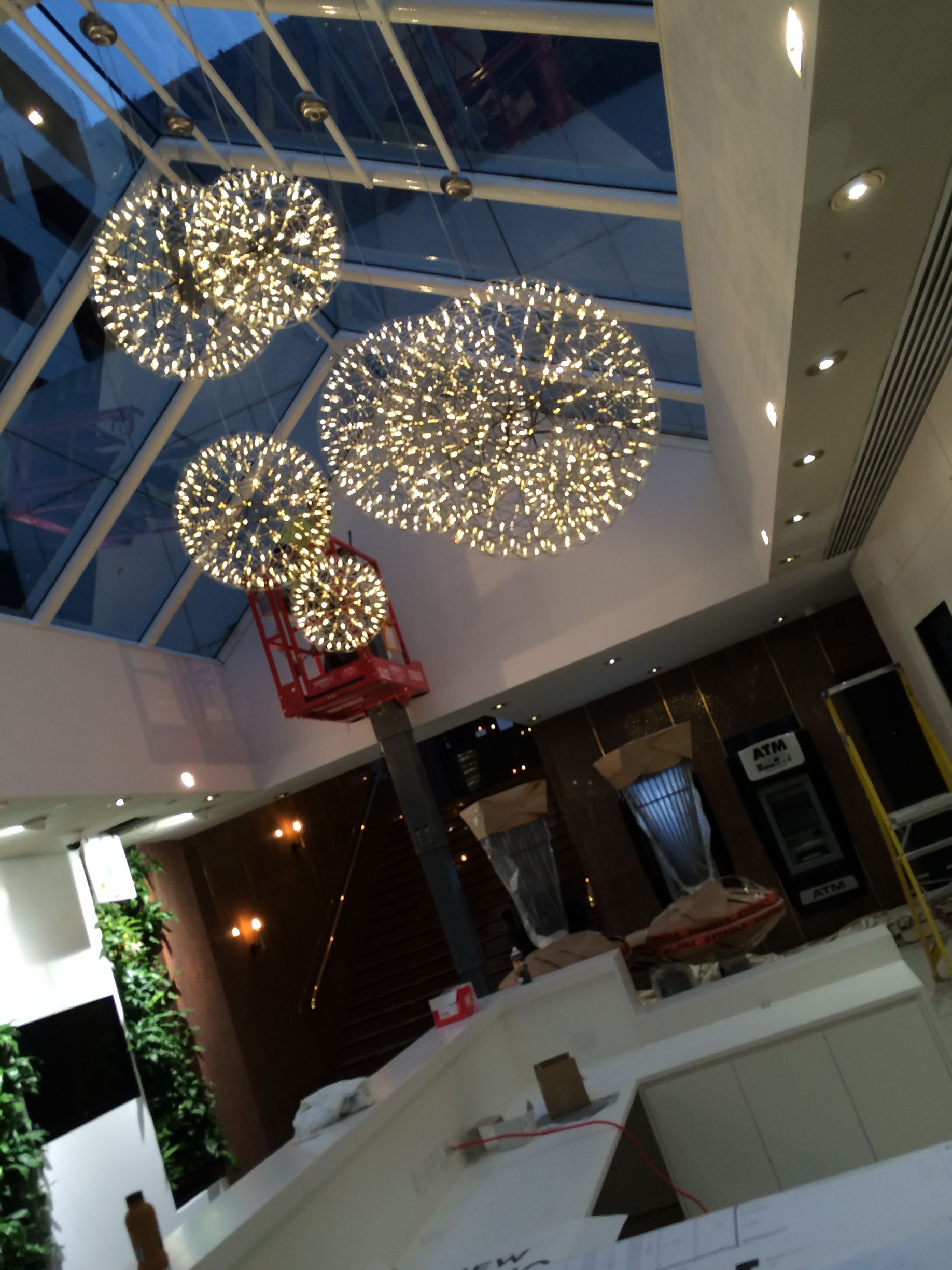 Four illuminated sphere chandeliers hanging from a glass ceiling; a person on a lift appears to be working on them.
