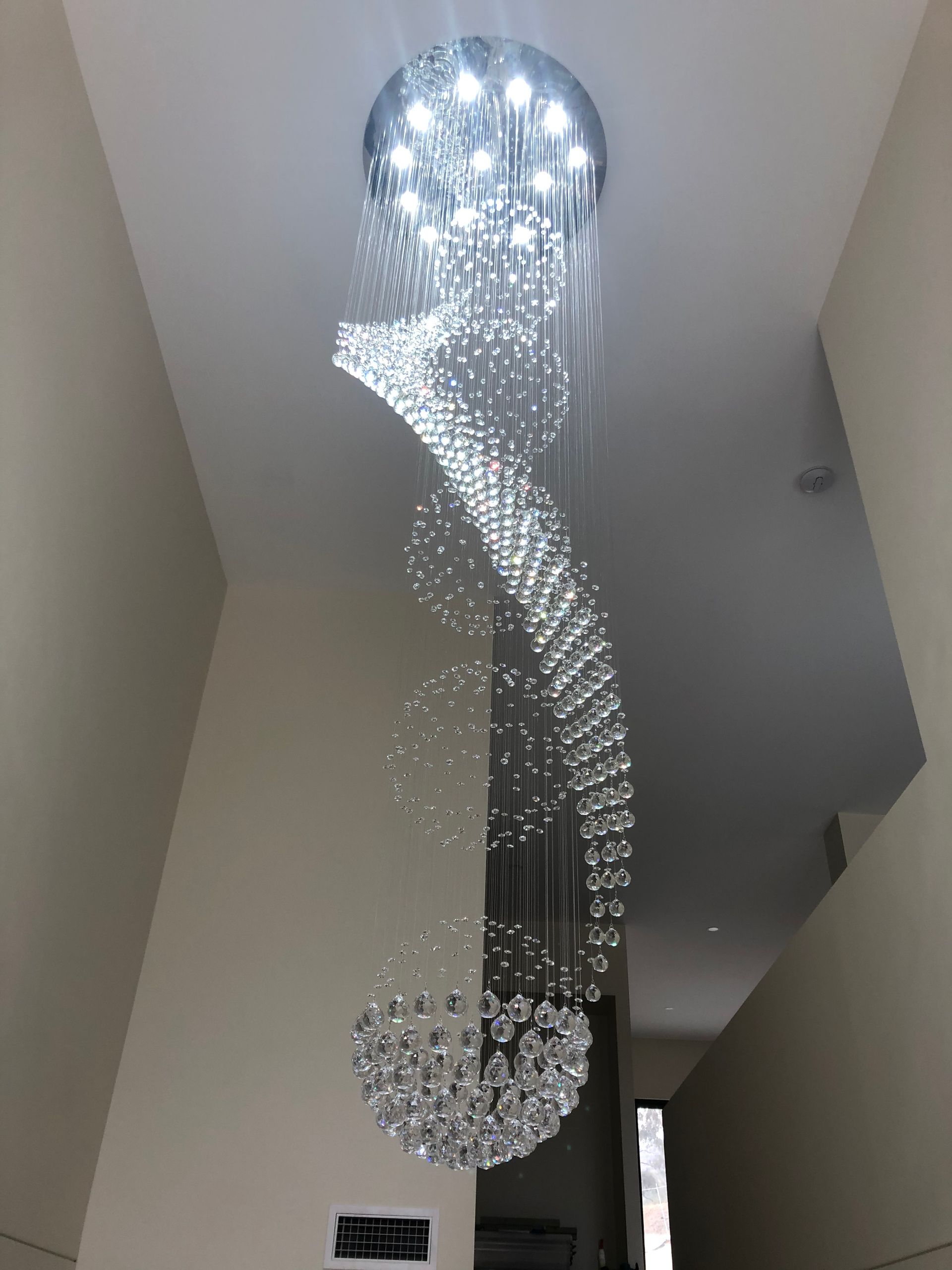 A large, spiral crystal chandelier hangs from a high ceiling, casting a brilliant light in a neutral-toned hallway.