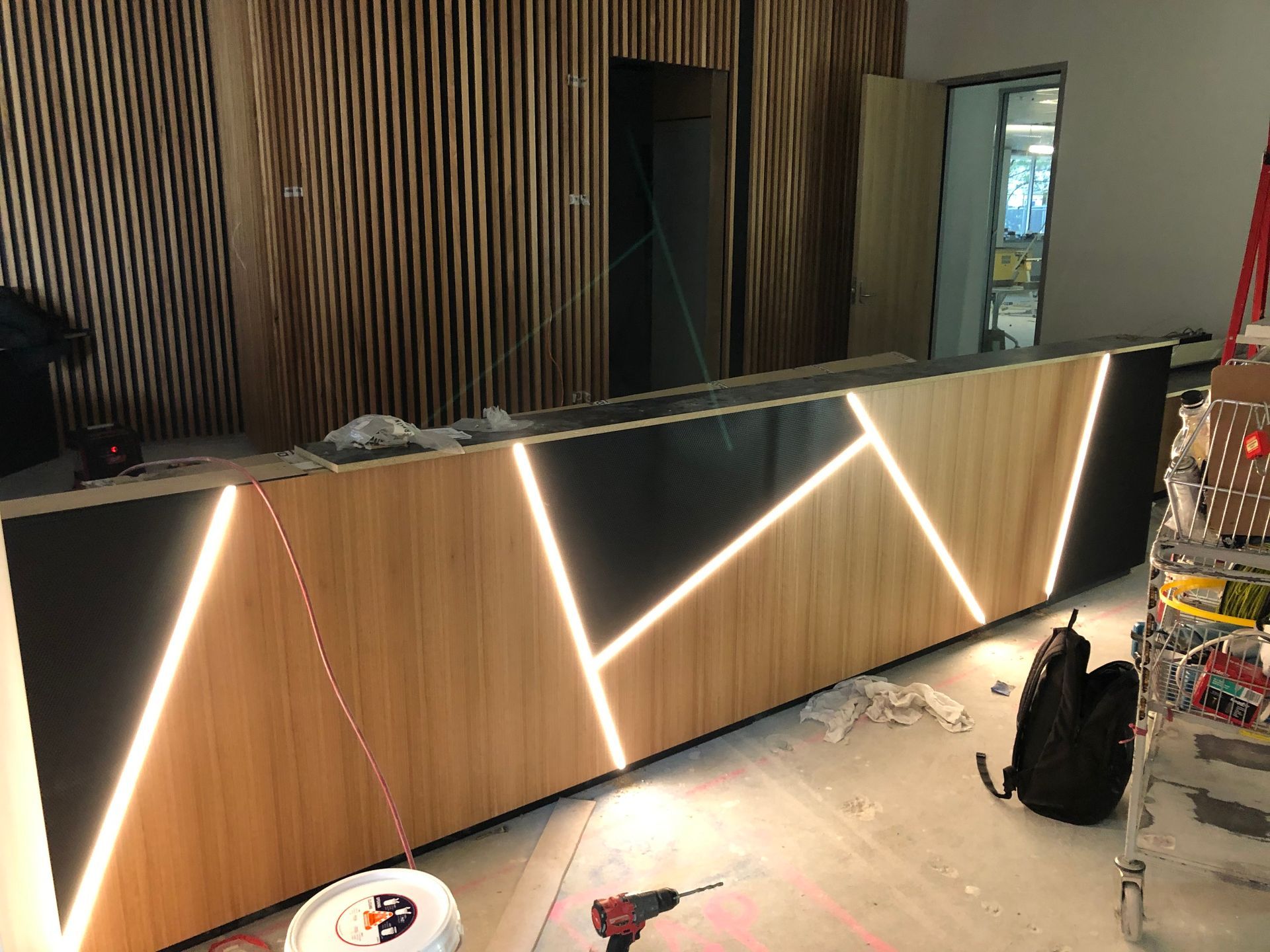 A modern reception desk with wooden and black panels, accented by glowing white geometric lines, under construction in a light-filled room.