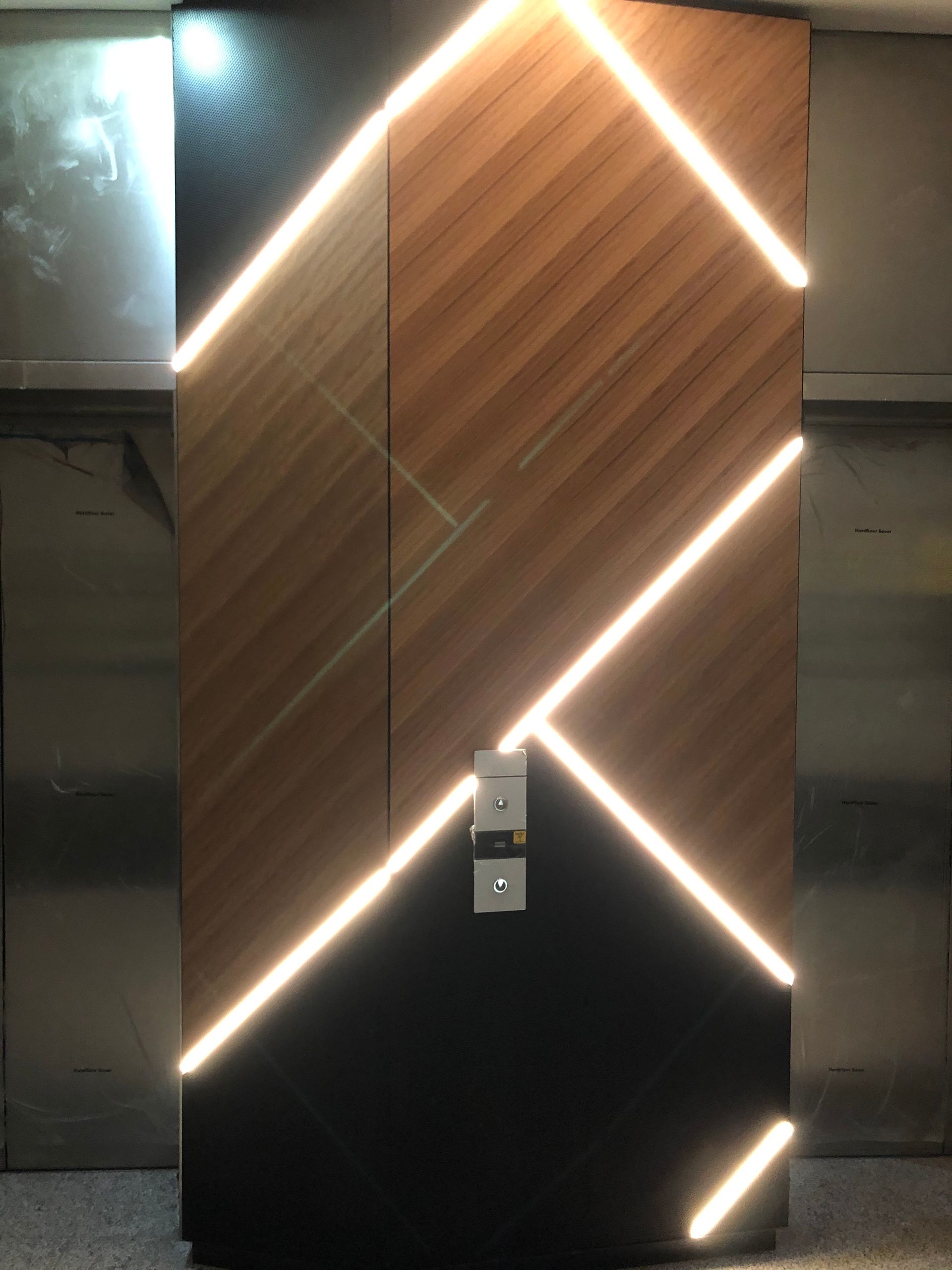 Modern wooden door with diagonal LED light accents, a black lower section, and a metal handle. It’s set in a metallic elevator