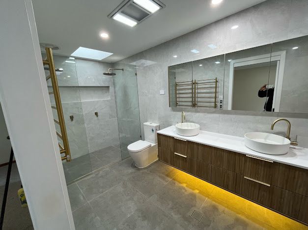 Modern bathroom with double vanity and walk-in shower; gold fixtures and trim; wood cabinetry.