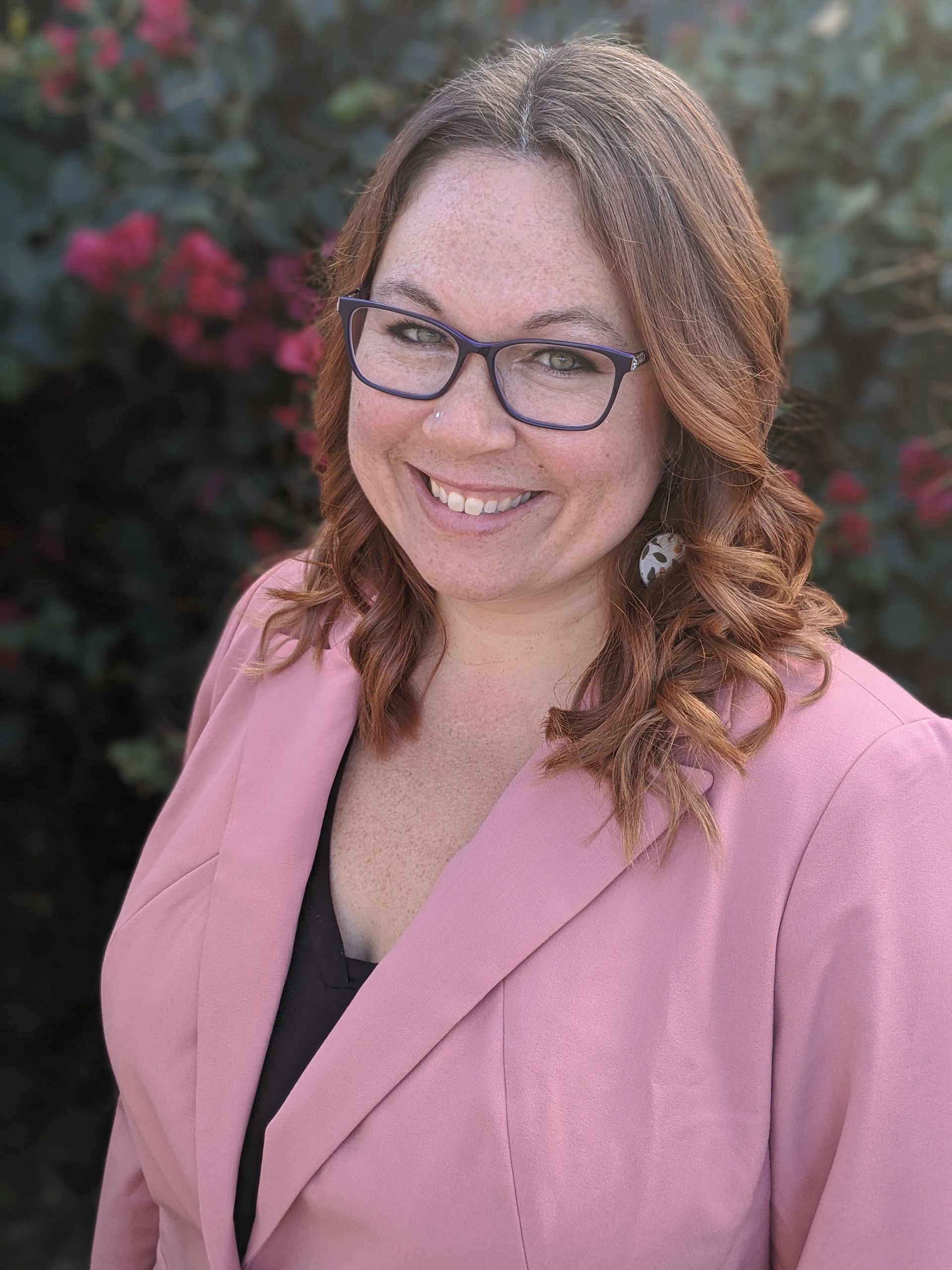 A woman wearing glasses and a pink jacket is smiling for the camera.