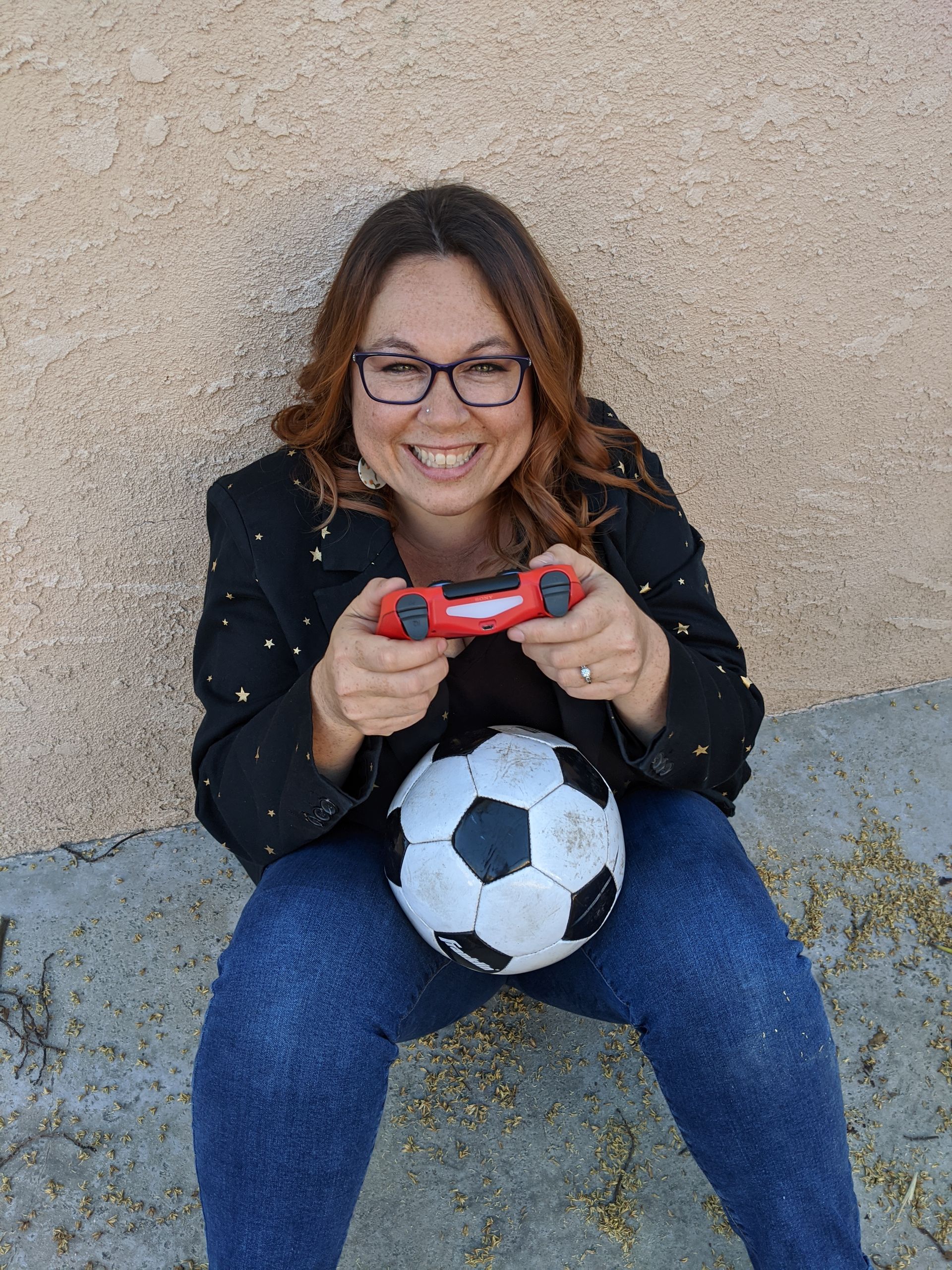 A woman is sitting on the ground holding a video game controller and a soccer ball.