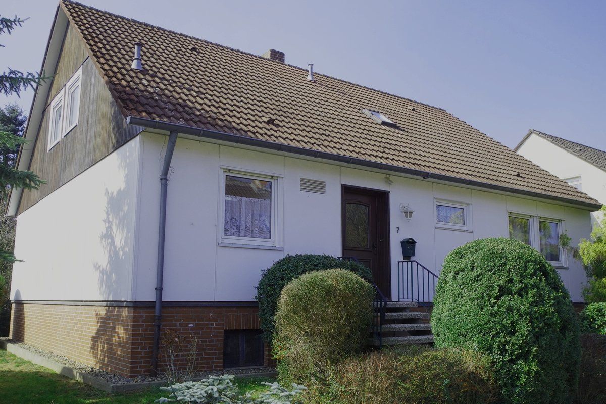 Ein weißes Haus mit braunem Dach ist von Büschen und Bäumen umgeben.