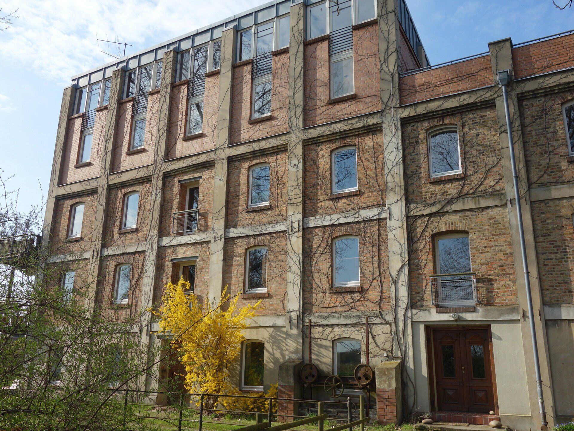 Ein großes Backsteingebäude mit vielen Fenstern und einem gelben Baum davor.