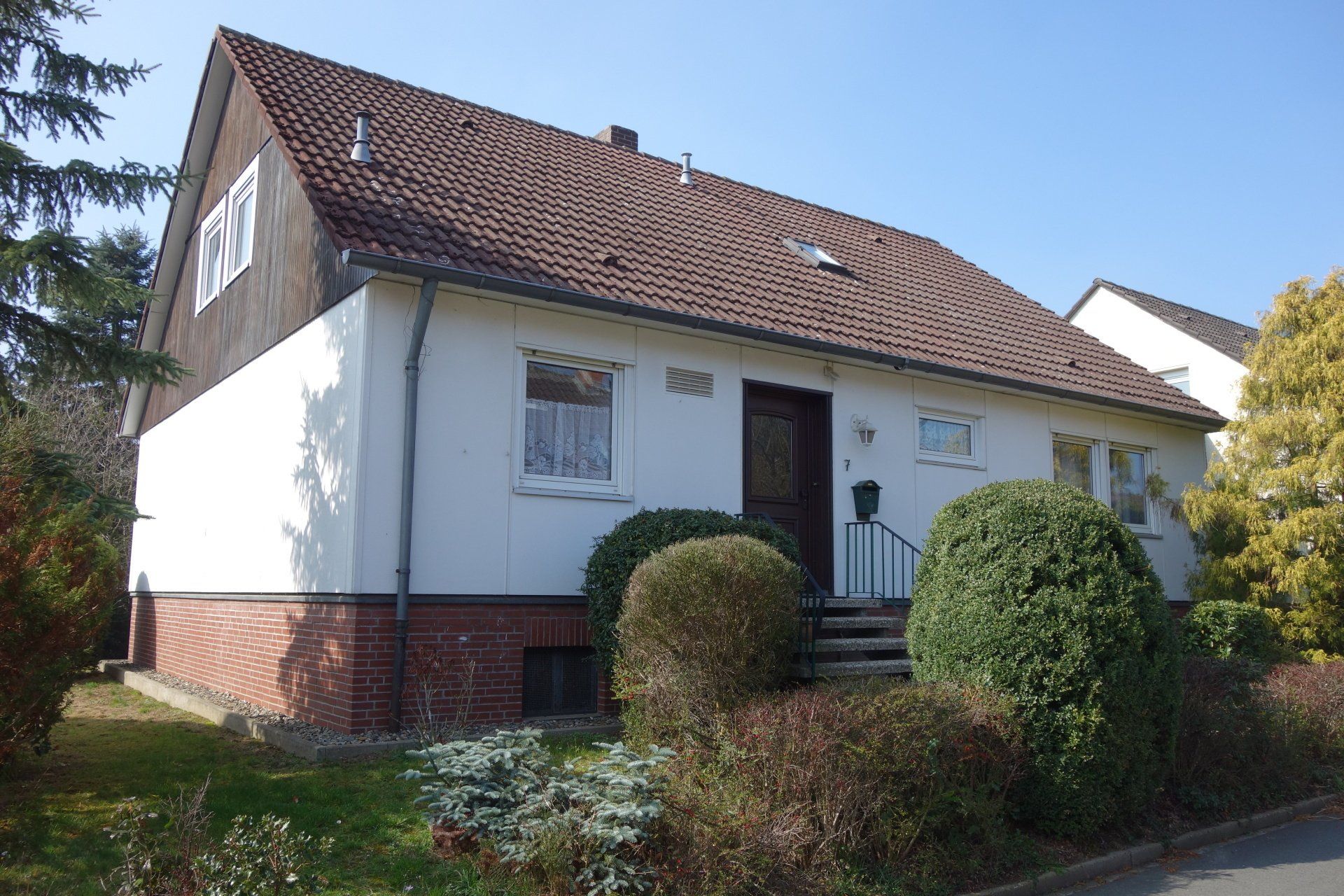 Ein weißes Haus mit einem braunen Dach ist von Büschen und Bäumen umgeben