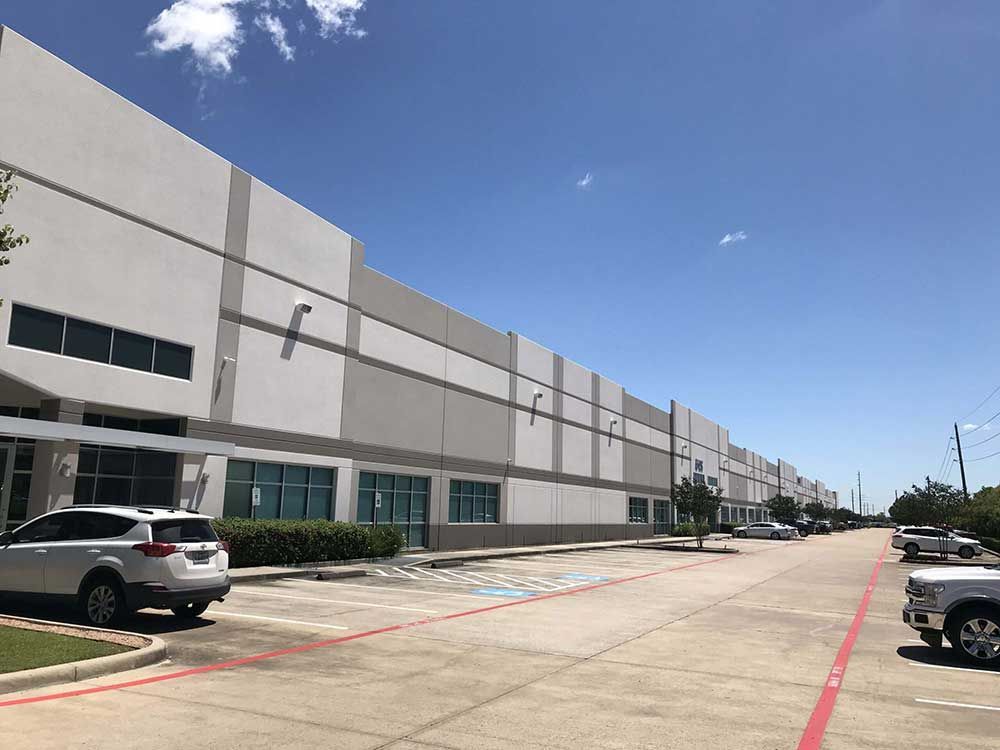 Long industrial building with gray and white panels, blue sky, parked cars.