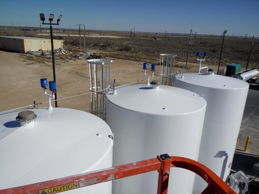 Three white storage tanks outdoors on a sunny day with blue devices on top, next to a brown, arid landscape.