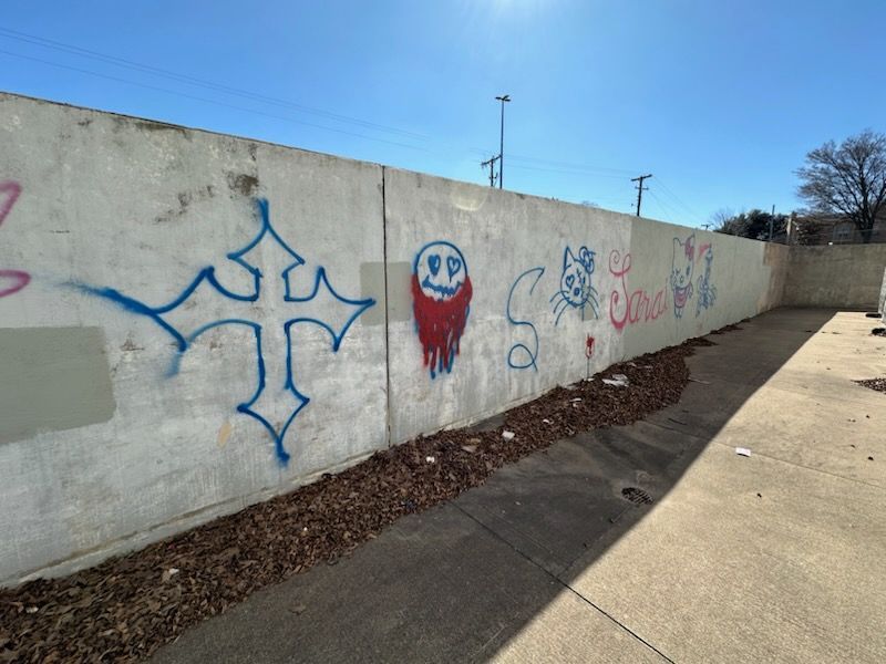 Concrete wall with graffiti art: cross, face, cat, text. Dry leaves and blue sky.