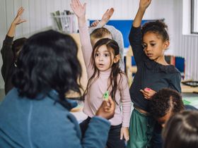 A group of children is raising their hands to answer a question in a classroom.