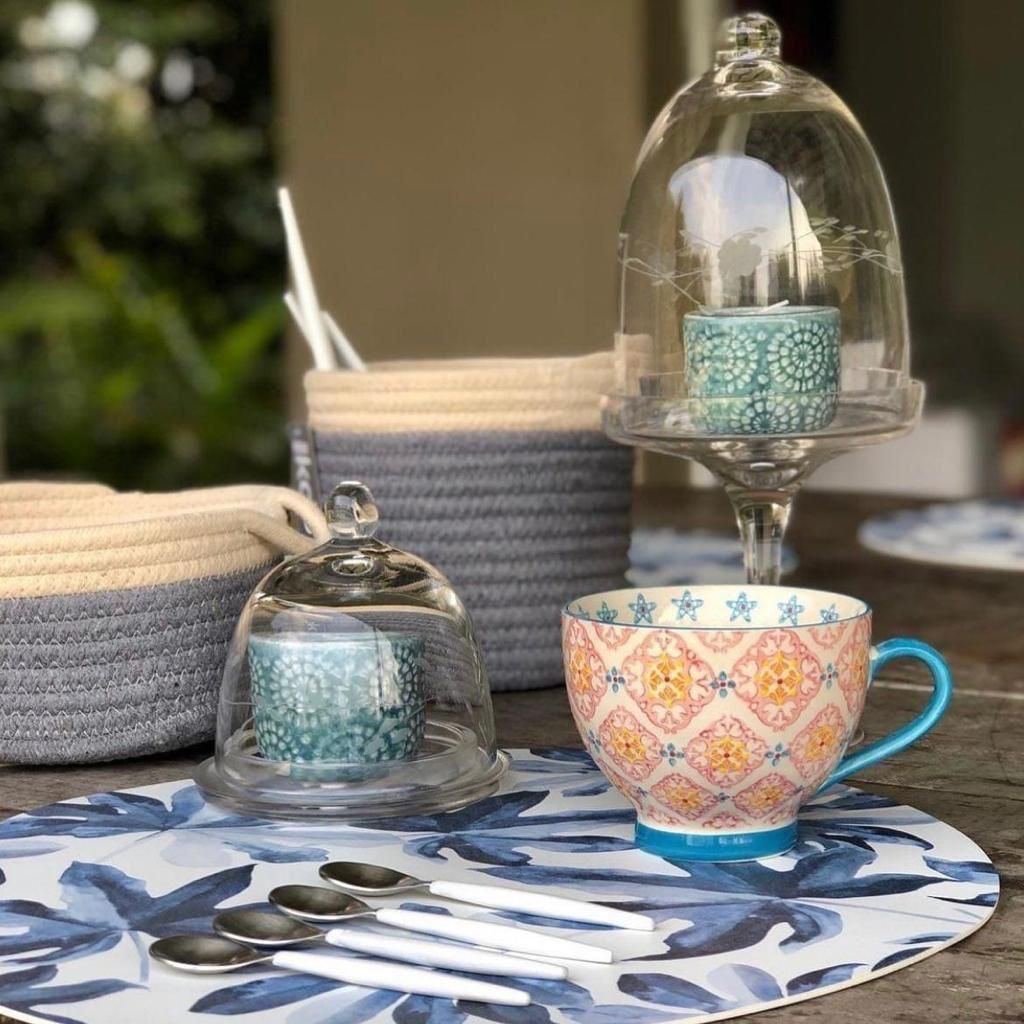 Configuración de mesa con mantel floral, taza estampada, campanas de cristal, cestas y cucharas.