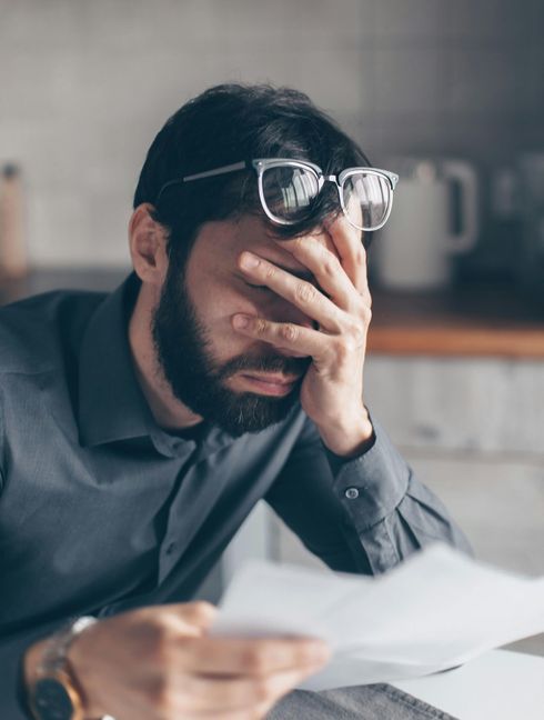 A man wearing glasses is covering his face while looking at a piece of paper.