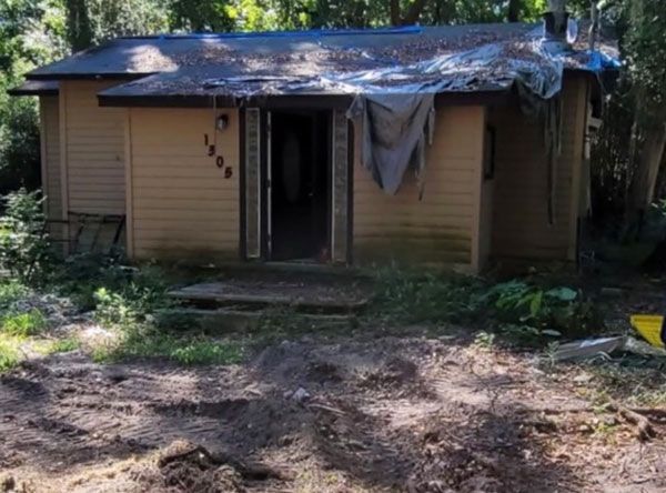 A small house with a tarp on the roof is surrounded by trees and dirt.