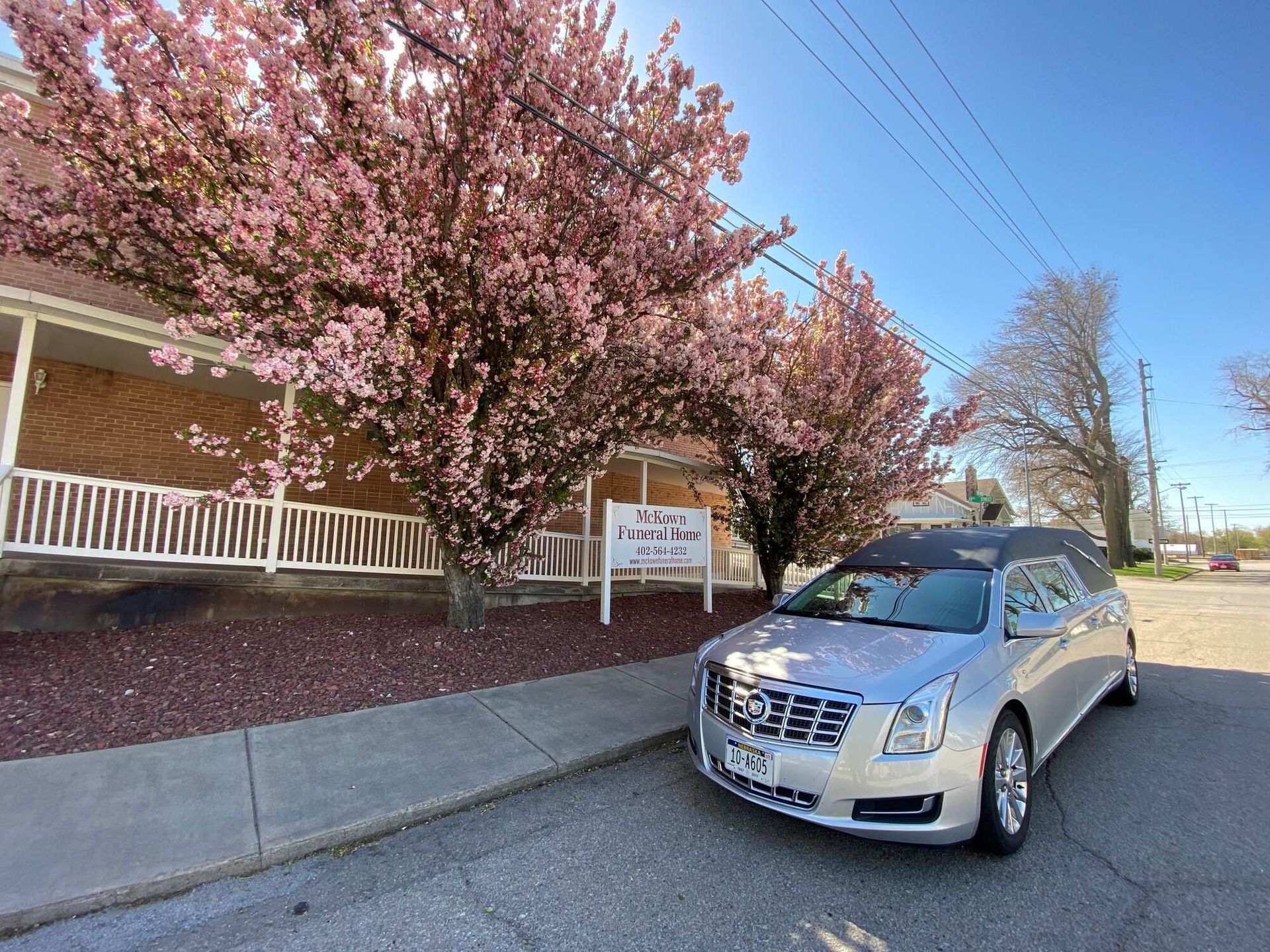 Silver hearse parked in front of a funeral home with blooming pink trees under a blue sky.