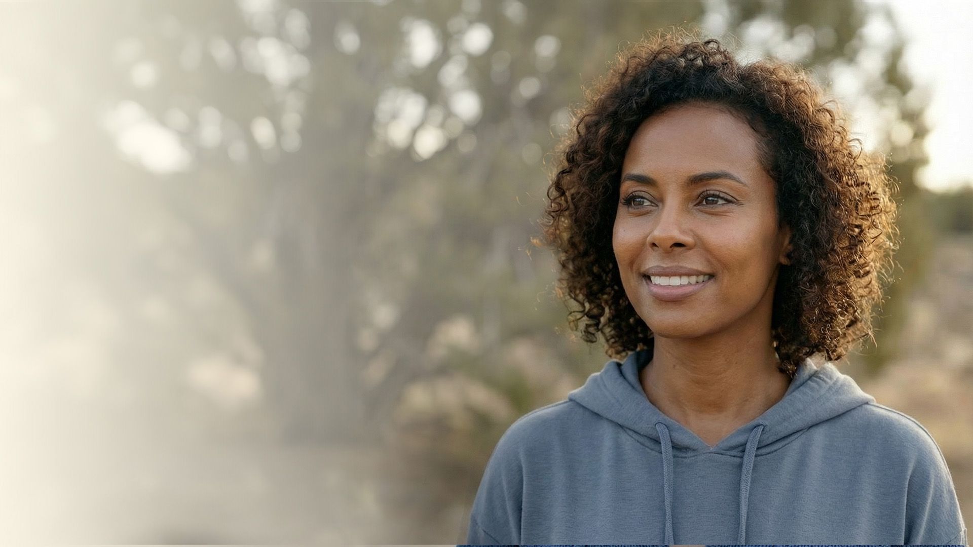 Woman with curly hair smiles outdoors, wearing a blue hoodie, looking toward the right.